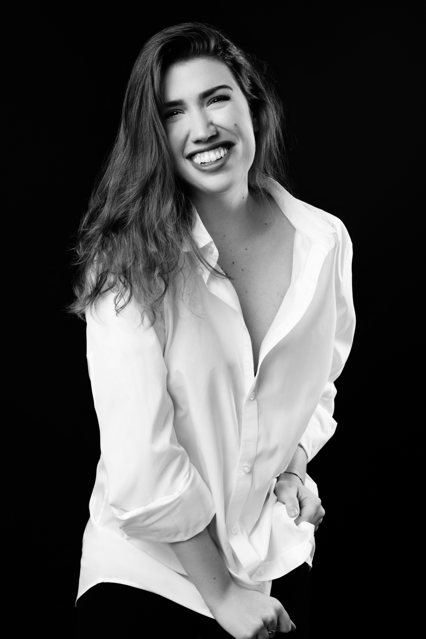 Black and white portrait of a young woman smiling broadly, wearing a white button-up shirt with rolled sleeves against a black background.