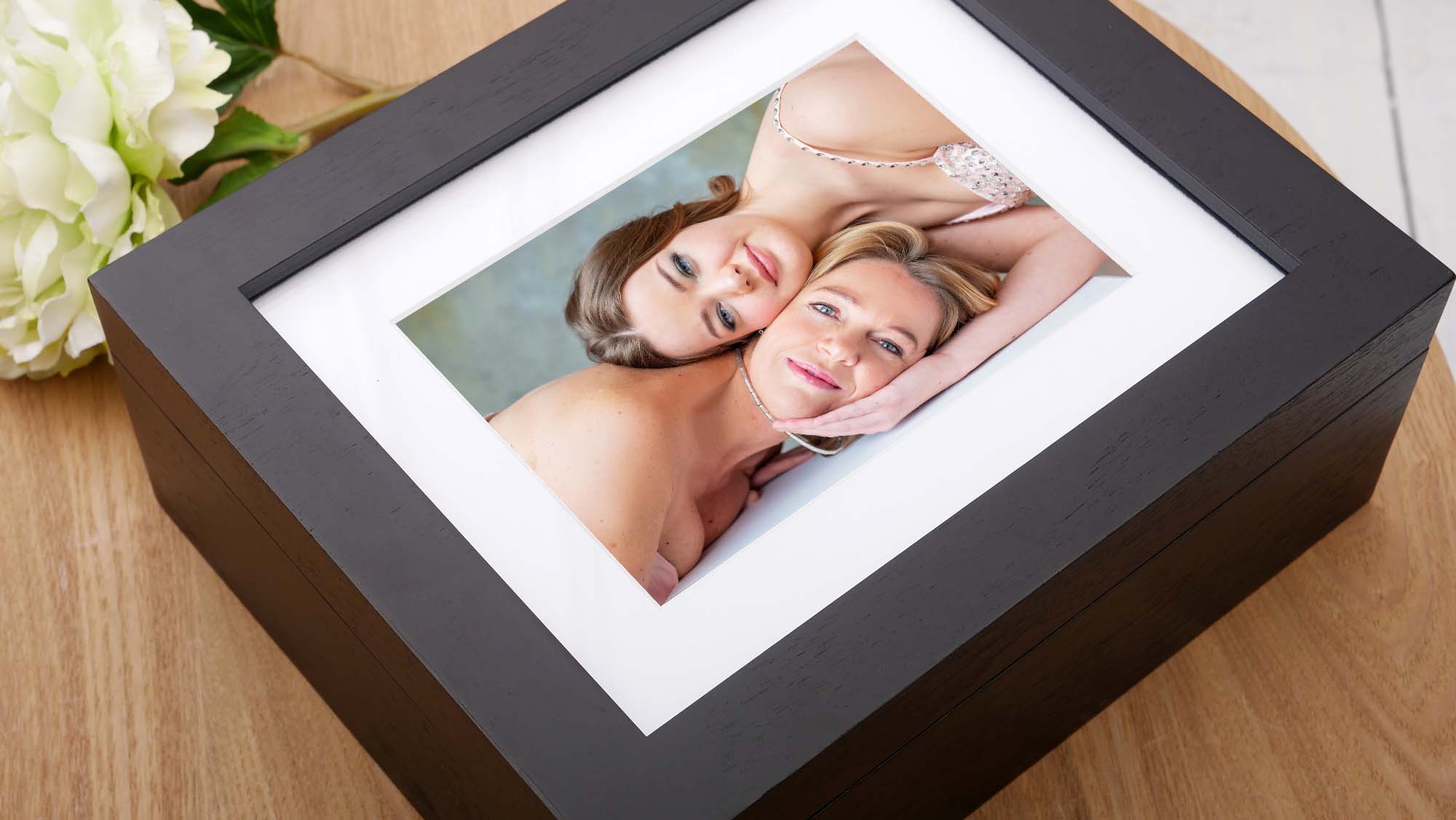 Black photo box on a wooden surface with a framed portrait of two women, one resting her head on the other's shoulder, both smiling gently.