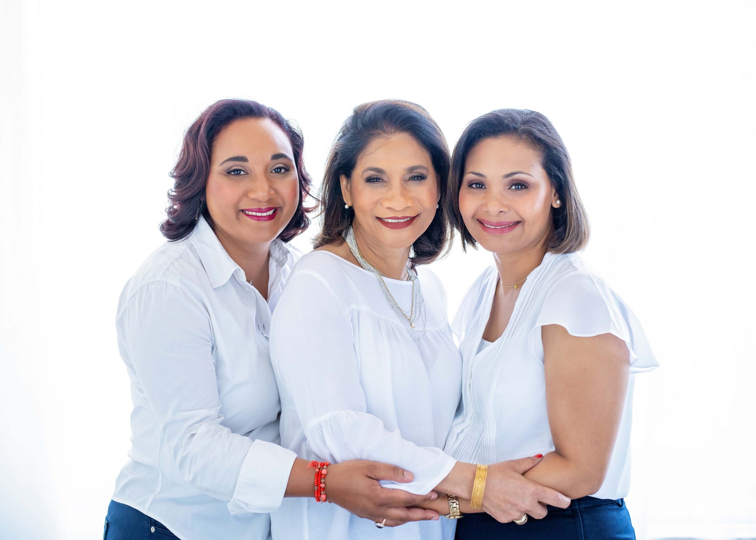 Three women standing close together, smiling and embracing each other, all wearing white tops against a bright white background.