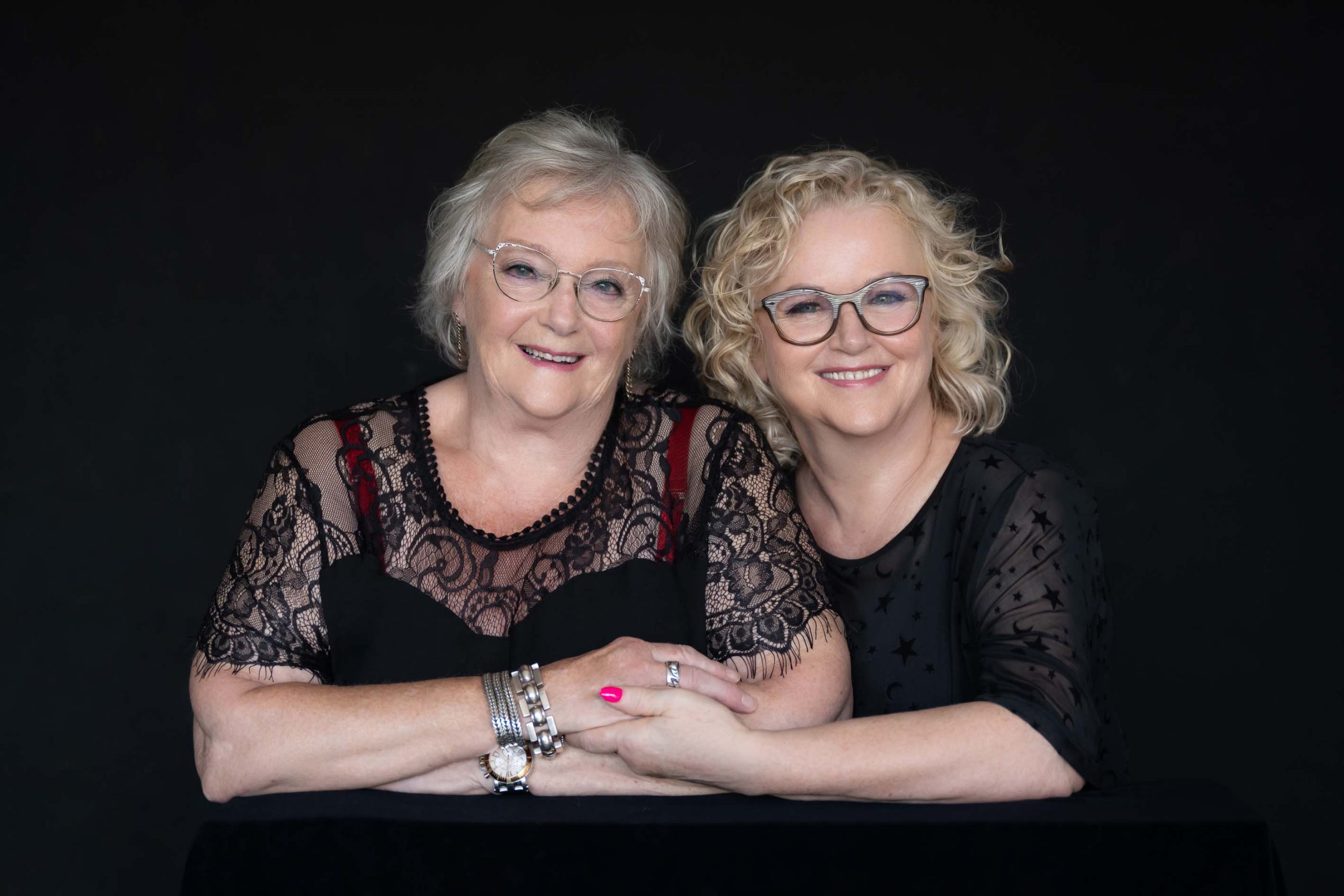 Portrait of two smiling women wearing glasses and black lace tops, sitting closely together with arms intertwined against a black background.