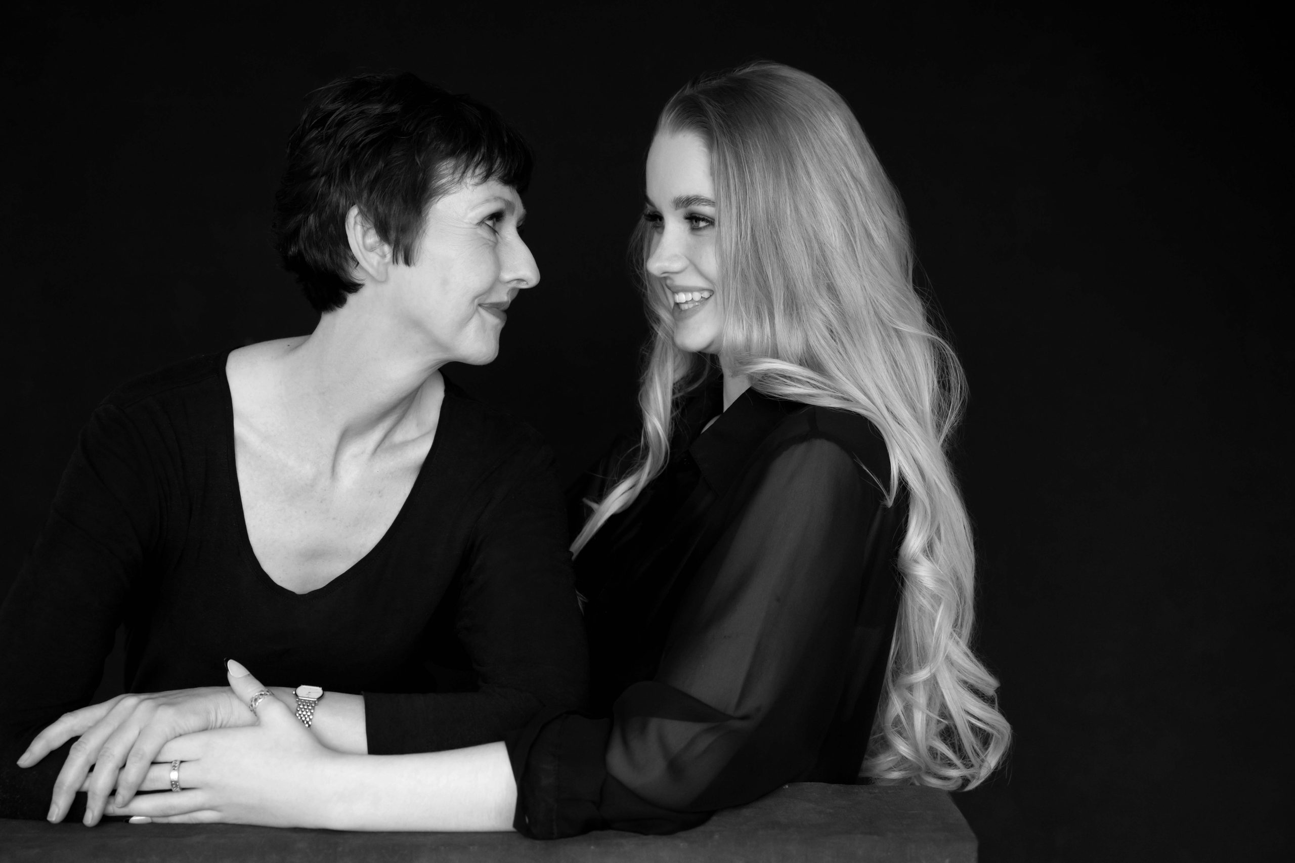 Black and white portrait of two women facing each other and smiling, with their arms gently intertwined on a surface between them against a dark background.