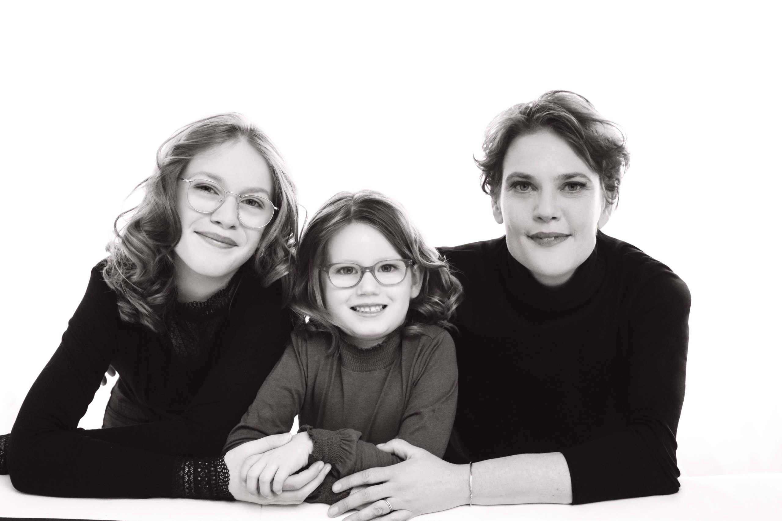 Black and white portrait of three females smiling and leaning forward with their arms crossed on a surface, featuring an adult woman on the right, a young girl in the middle, and an older girl on the left, all wearing glasses and dark tops.