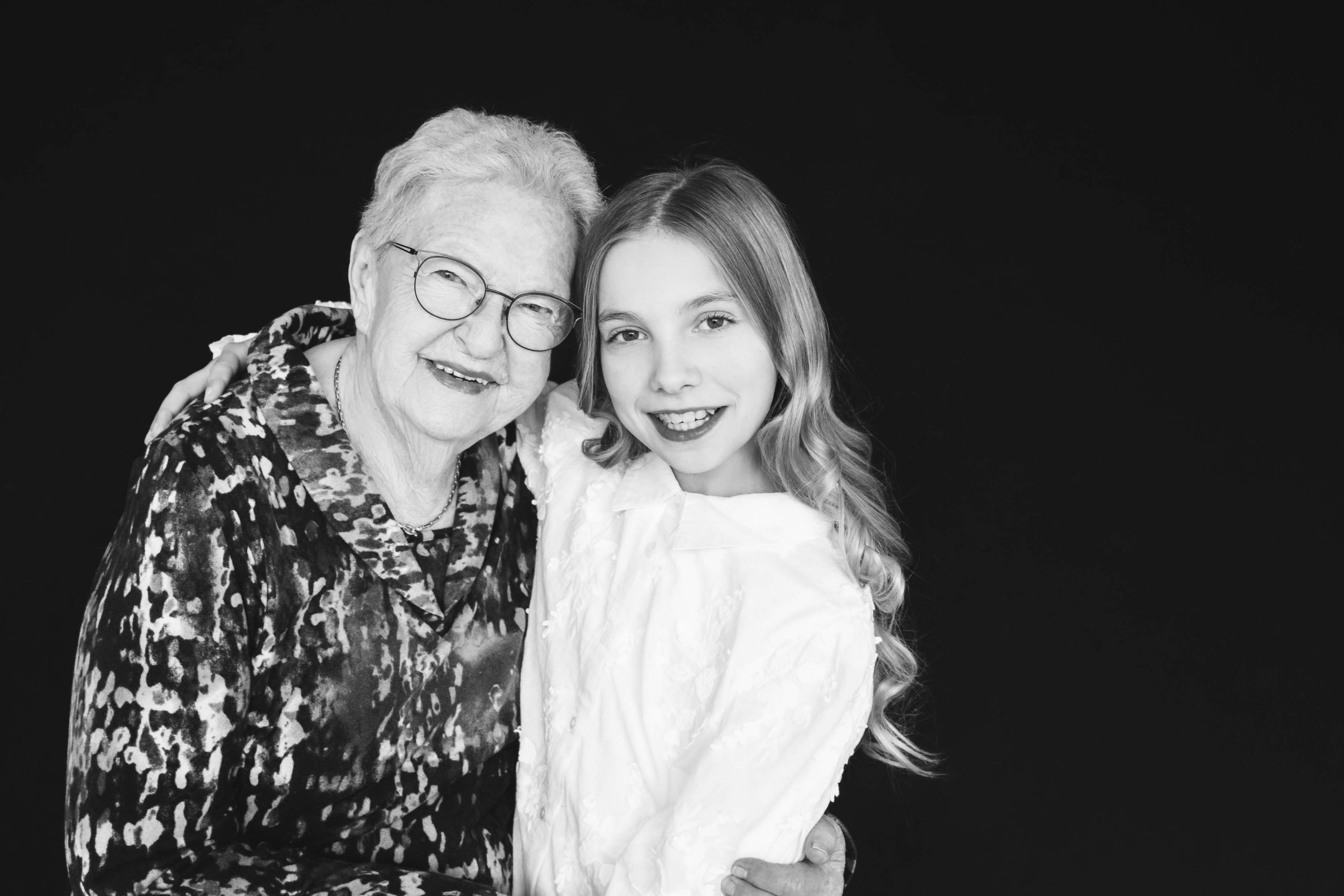 Black and white photo of an elderly woman with glasses and a patterned blouse embracing a smiling young girl with long wavy hair wearing a white textured blouse, both looking at the camera against a black background.