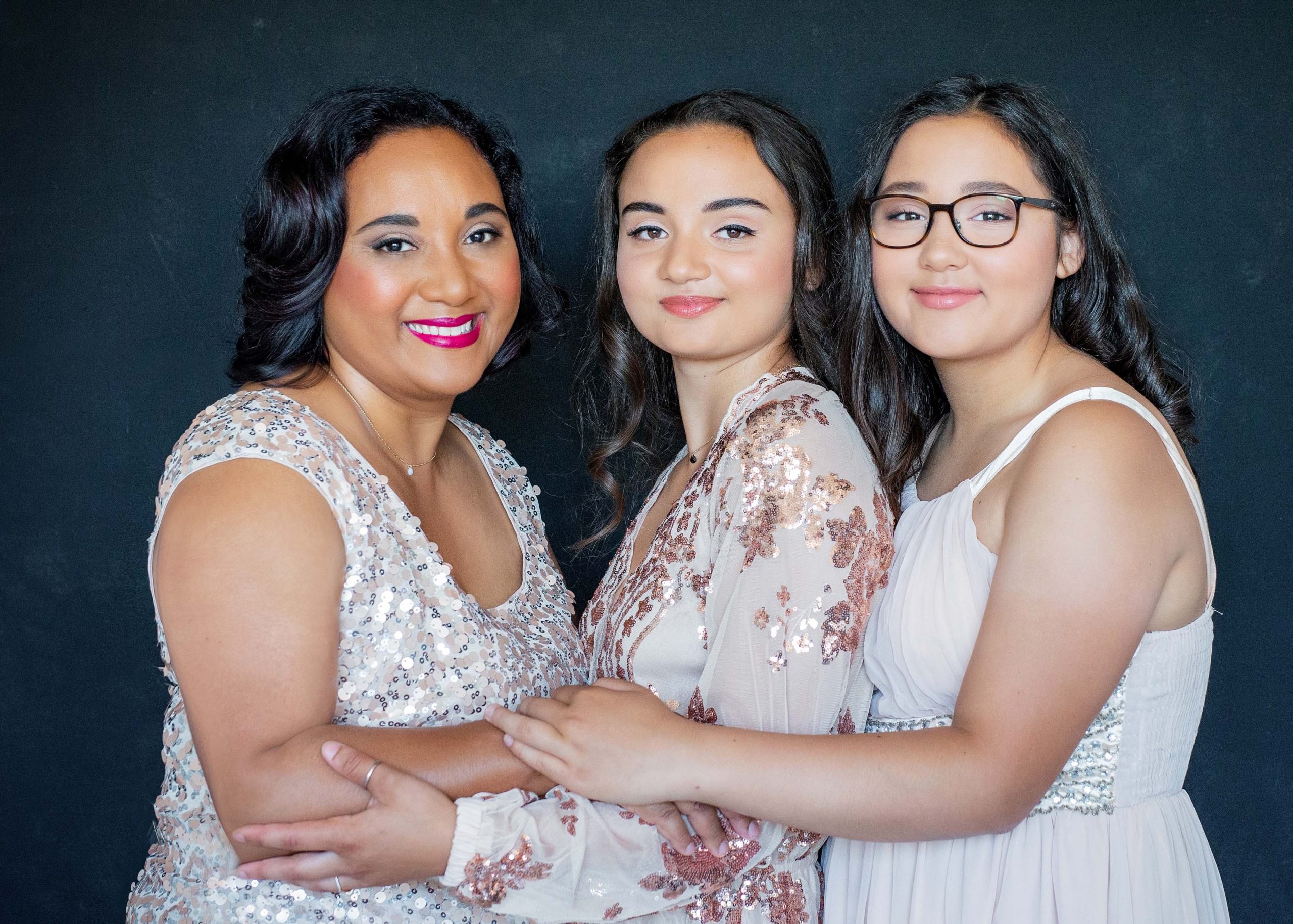 Three women in fancy dresses standing together against a dark background, smiling warmly and embracing each other closely.