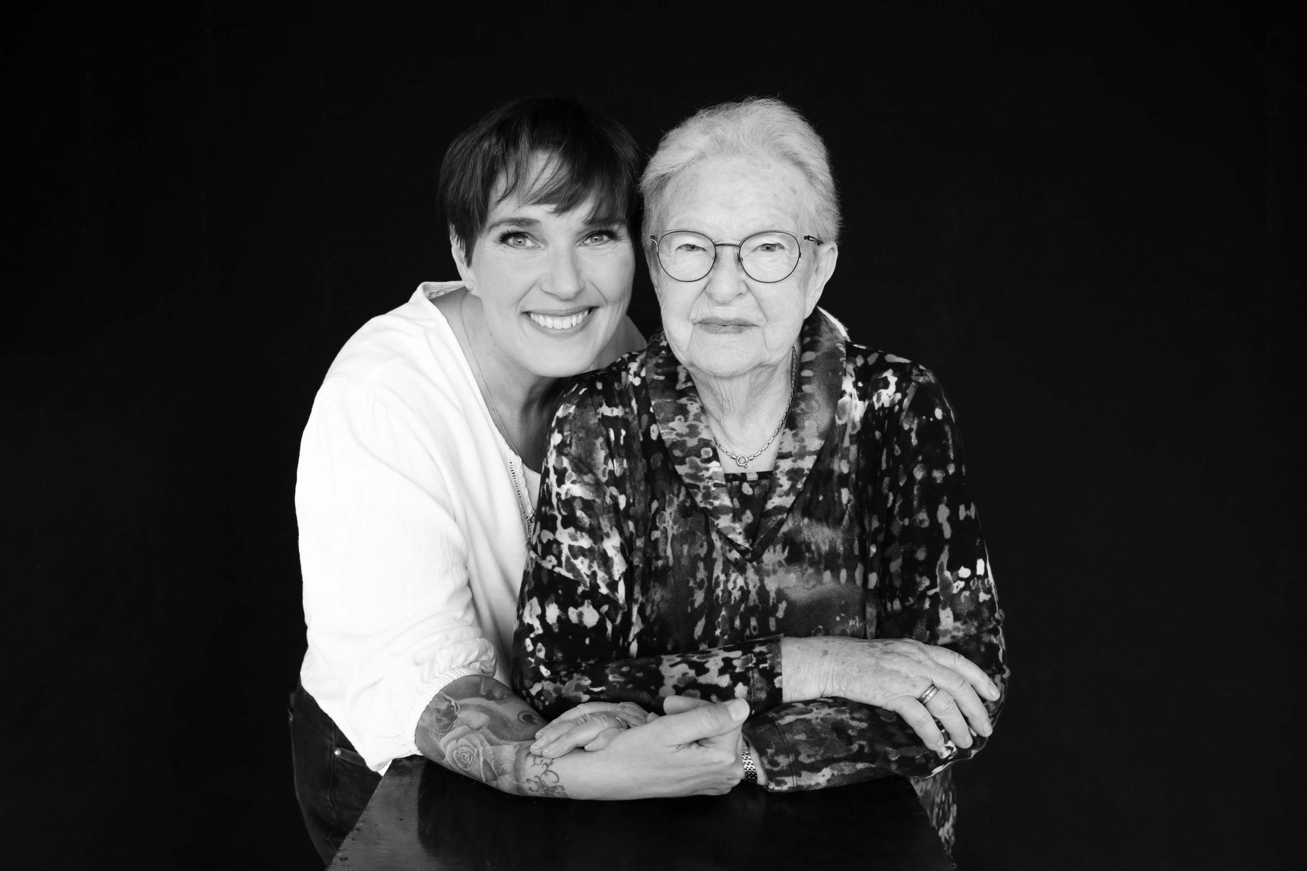 Black and white portrait of a smiling middle-aged woman with a tattoo on her arm embracing an elderly woman wearing glasses and a patterned blouse, both sitting at a table against a black background.