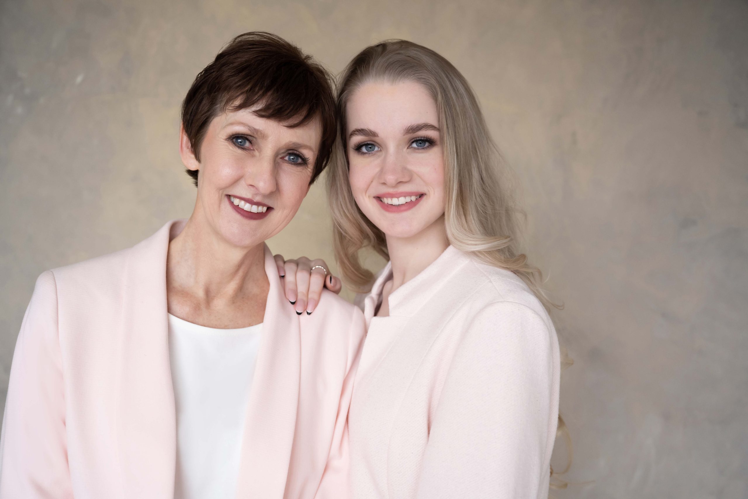 Smiling middle-aged woman with short brown hair and young woman with long blonde hair standing close together, both wearing light pink jackets, on a neutral background.