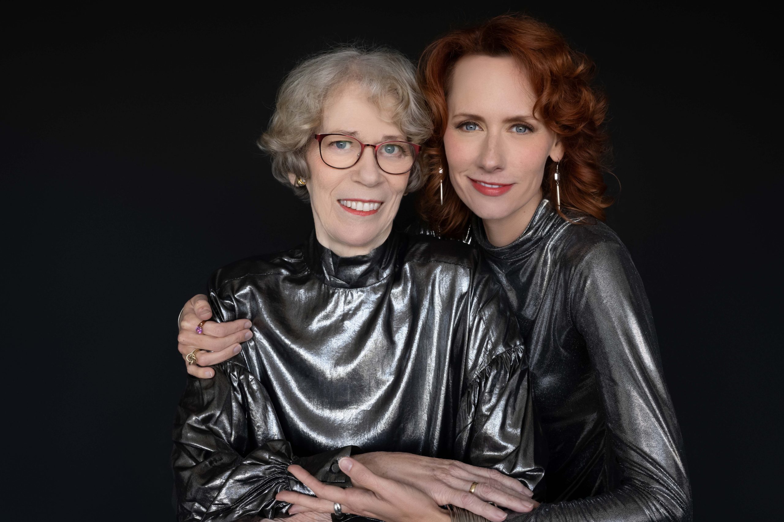 An older woman with short gray hair and glasses embraces a younger red-haired woman, both wearing shiny metallic silver tops, smiling against a black background.