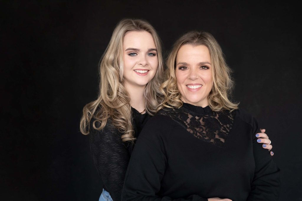 A smiling woman with blonde curly hair wearing a black lace top is embraced from behind by a younger woman with similar blonde curly hair wearing a black long sleeve shirt and light blue jeans, against a black background.