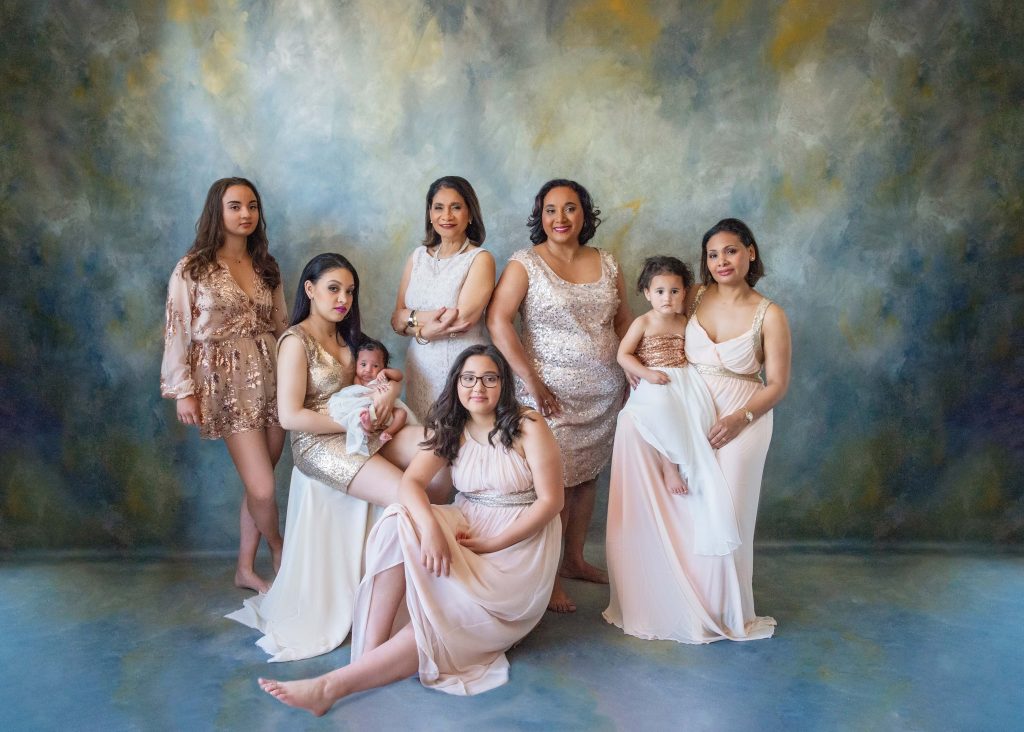Seven females of different ages dressed in semi-formal and formal dresses posing against a painted blue and gold backdrop; one woman is seated holding a baby, another child is standing on a woman’s long dress, and another woman is seated on the floor.