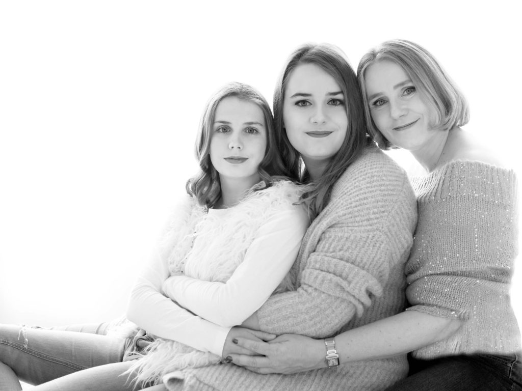 Black and white portrait of three women sitting closely together, smiling gently, with the woman on the right wearing a knit sweater, the middle woman wearing a cozy cardigan, and the woman on the left wearing a furry vest over a long-sleeve shirt.