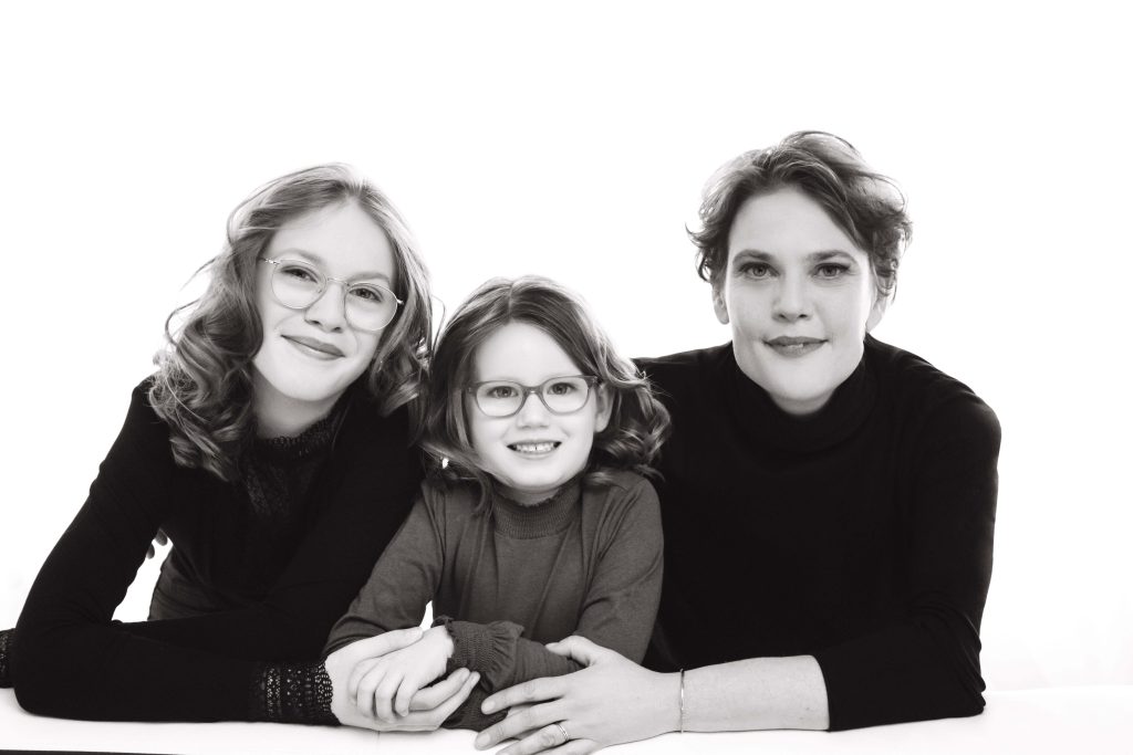 Black-and-white portrait of a woman and two girls with curly hair and glasses, all smiling and resting their arms on a white surface.