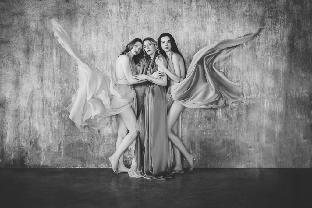 Black and white photo of three women standing barefoot against a textured wall, wearing flowing dresses with fabric lifted to resemble wings, posing closely together and looking at the camera.