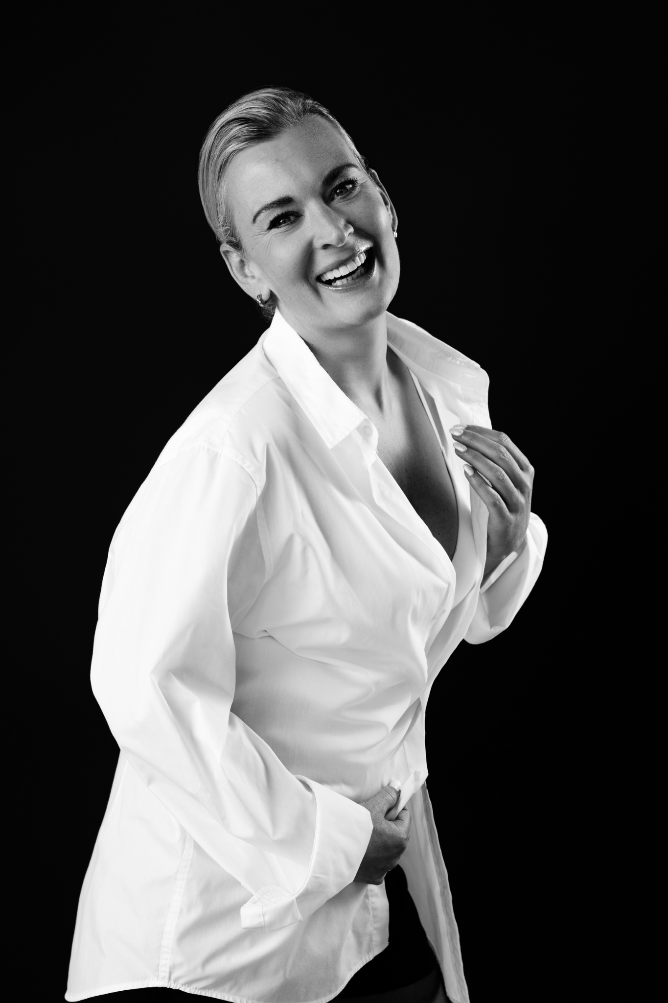 Black and white portrait of a smiling woman with short hair wearing a white button-up shirt against a black background.