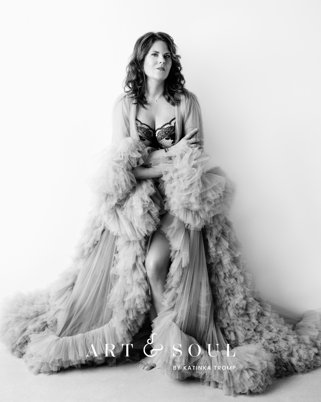 Black and white photo of a woman standing barefoot against a white background, wearing a lace bodysuit and a voluminous, ruffled tulle robe, with her arms crossed.