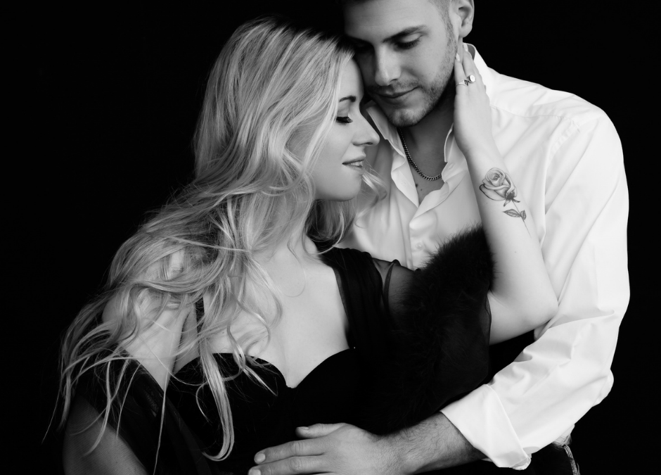 Black and white photo of a couple embracing closely; the woman with long hair and a rose tattoo on her forearm gently holds the man's face, while he looks down with a soft smile.