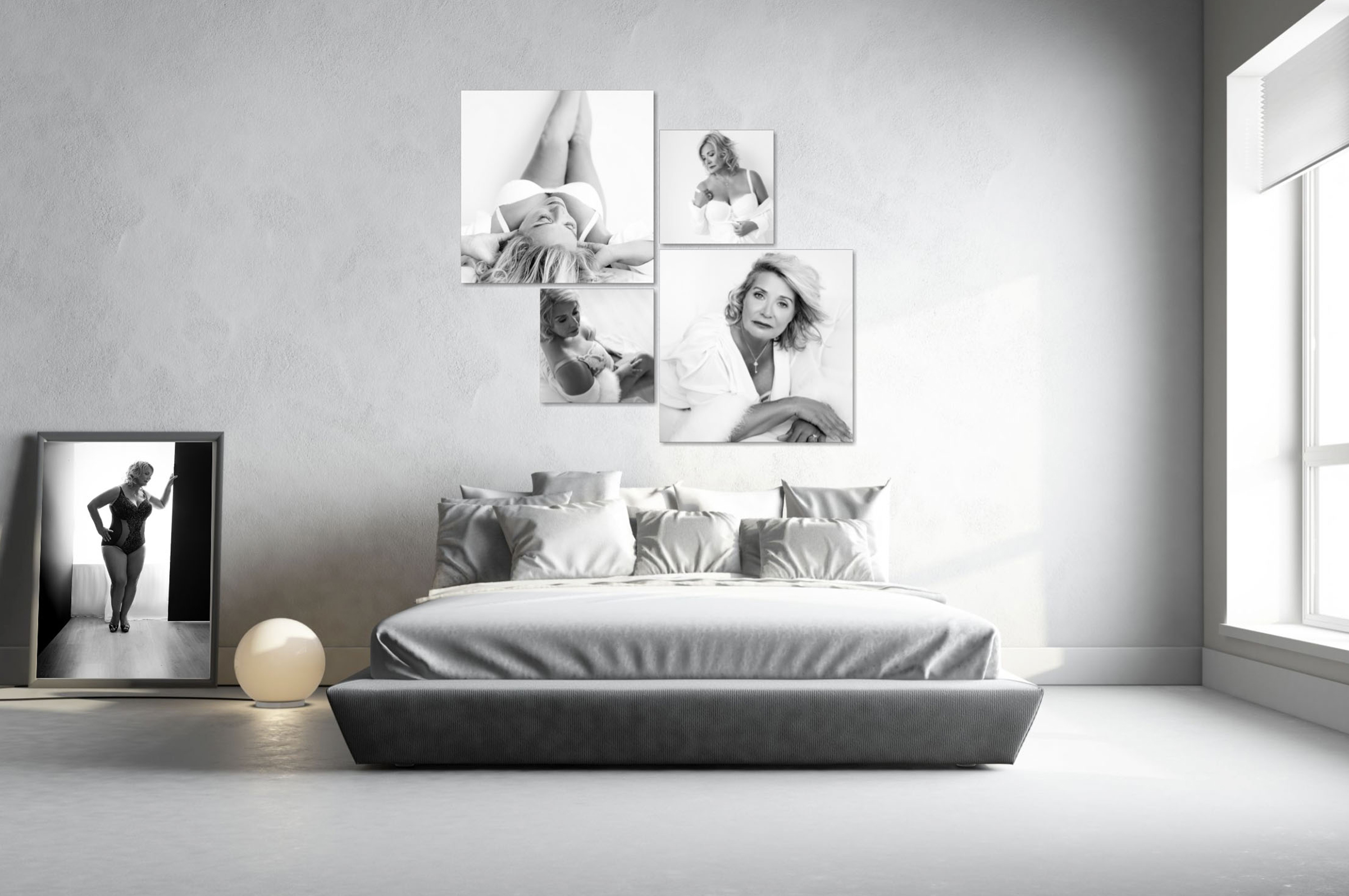 Minimalist bedroom with a large bed featuring multiple pillows and a padded headboard, illuminated by natural light from a window. On the wall above the bed are five black-and-white portraits of a woman in various poses. To the left, a large framed black-and-white photo of the same woman stands on the floor next to a round glowing lamp.