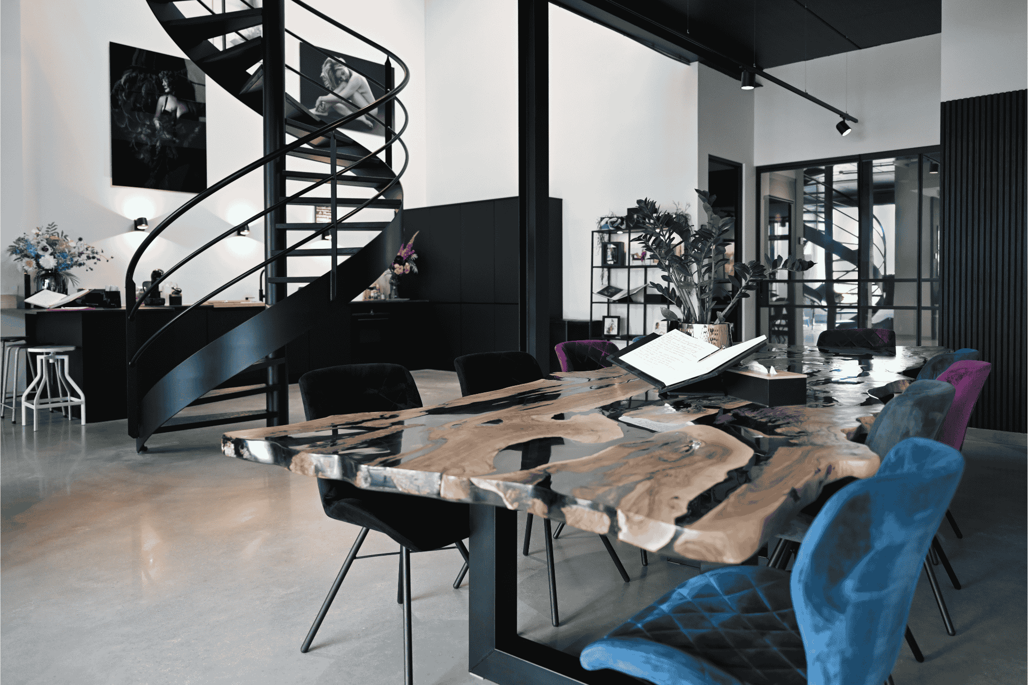 Modern interior featuring a large, artistic wooden and resin dining table surrounded by velvet chairs in blue, purple, and black, with a potted plant and open book on the table, and a black spiral staircase in the background against white walls.
