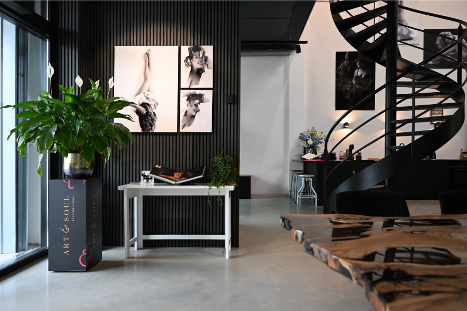 Modern art studio interior featuring a large leafy plant on a pedestal labeled 'Art & Soul,' a white table with photo books and a trailing plant, black-striped wall with black-and-white portrait photographs, a black spiral staircase leading to an upper level, and a live-edge wooden table in the foreground, all illuminated by natural and ambient light.
