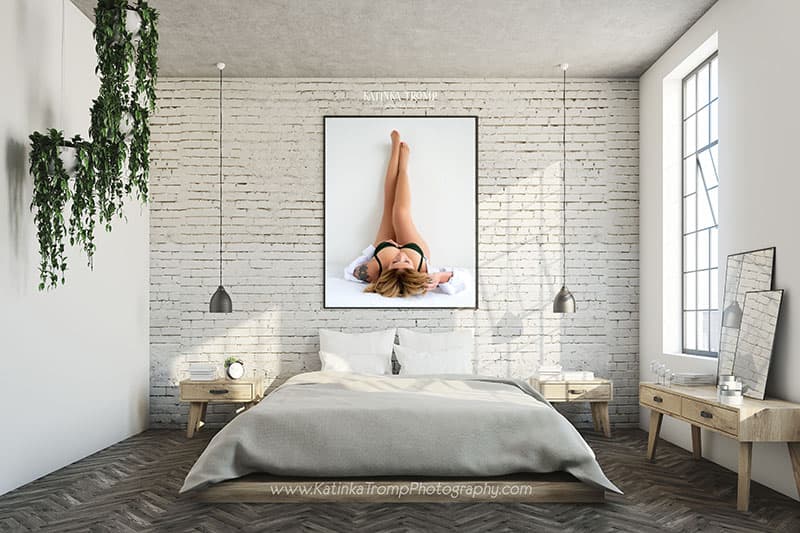 Modern minimalist bedroom with a low bed with white and gray bedding centered against a white brick wall, flanked by wooden nightstands with pendant lights above. Above the bed is a large framed photo of a woman lying down with legs raised against a white background. The room has a wooden floor, a large window on the right letting natural light in, hanging green plants on the left wall, and a wooden dresser with two leaning mirrors on the right.