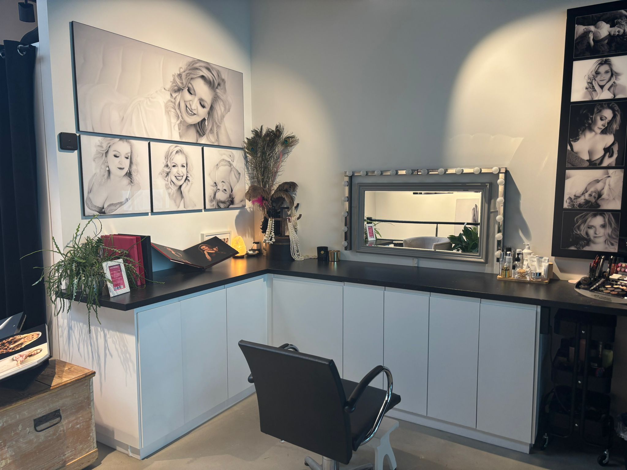 A stylish makeup or photography studio corner with a black swivel chair in front of a long mirror framed with round lights, surrounded by white cabinets and a variety of portrait photos of a smiling blonde woman on the walls, along with makeup products, a potted plant, decorative feathers, and a soft yellow light on the countertop.