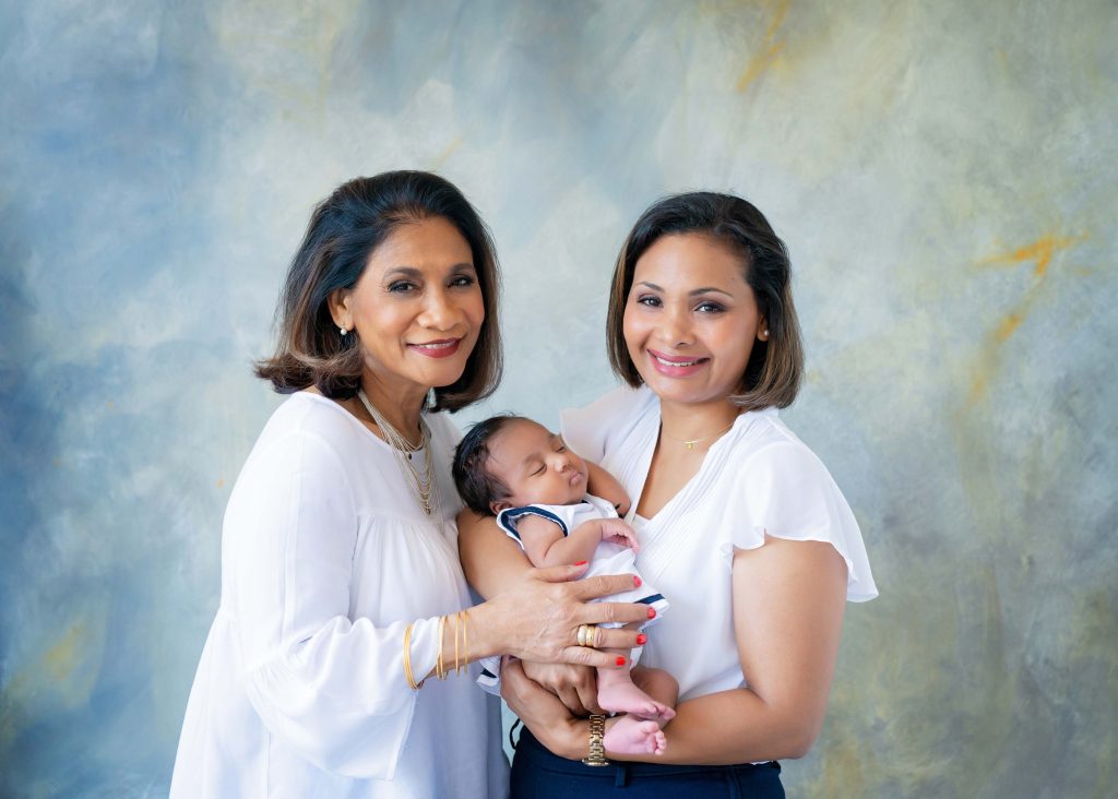 Two smiling women in white tops holding a sleeping baby dressed in white and navy blue, standing in front of a soft blue and yellow mottled backdrop.