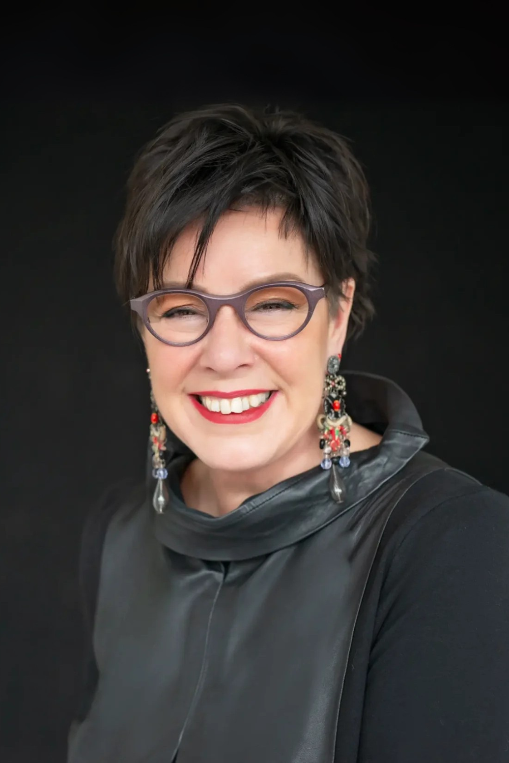 Smiling woman with short dark hair, wearing round glasses, red lipstick, intricate long earrings, and a black leather top against a black background.