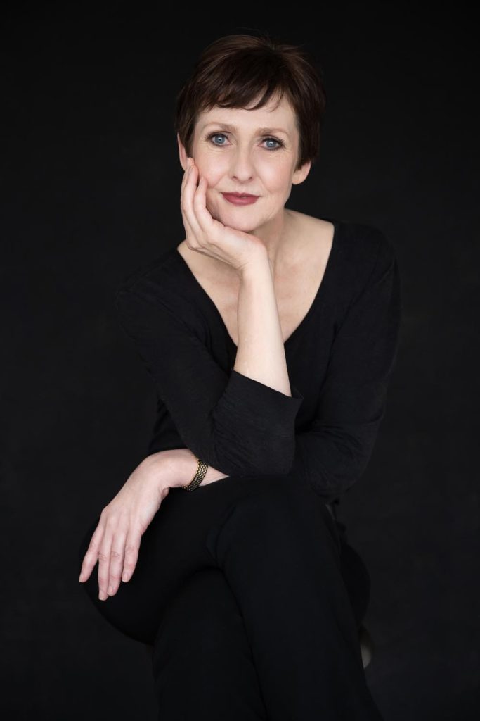 Portrait of a woman with short brown hair and blue eyes wearing a black top, resting her face on her hand against a black background.