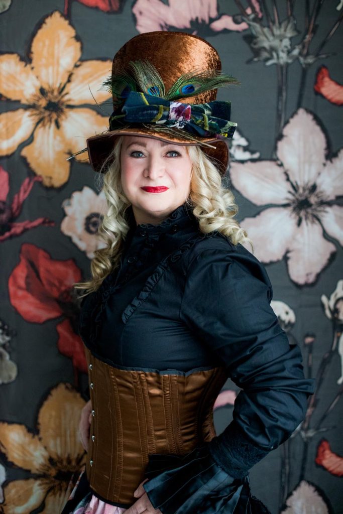 Woman with curled blonde hair wearing a tall brown velvet hat adorned with peacock feathers and a patterned bow, a black high-collared blouse, and a brown corset, standing against a floral background.