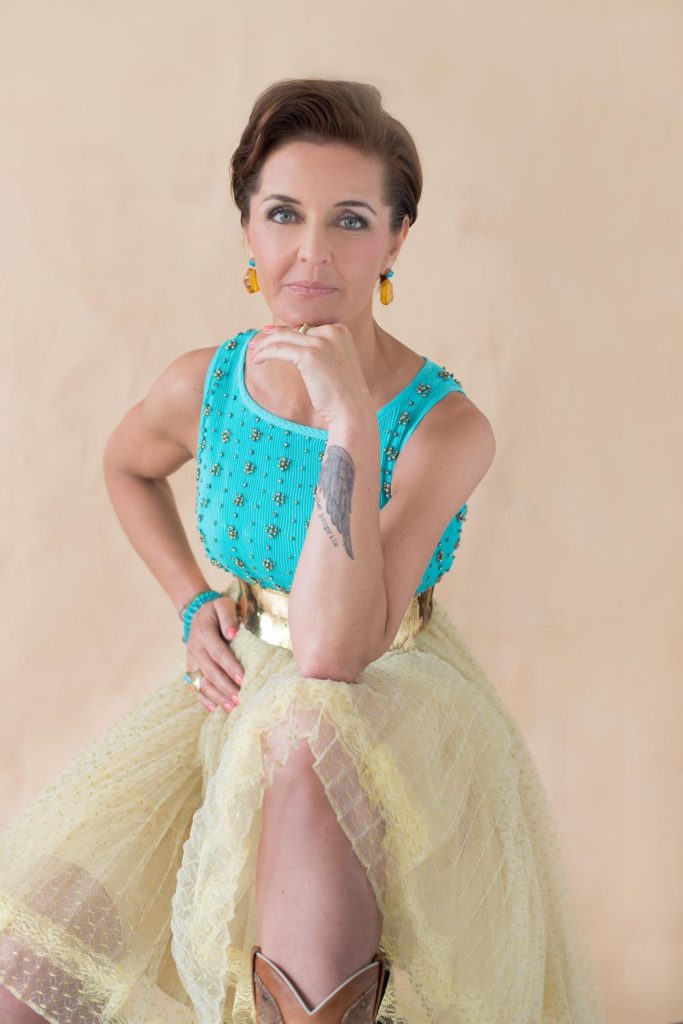 Woman with short brown hair wearing a turquoise sleeveless top with bead embellishments, a yellow tulle skirt, and brown boots, resting her chin on her hand, showing a tattoo on her forearm, posing against a plain light beige background.