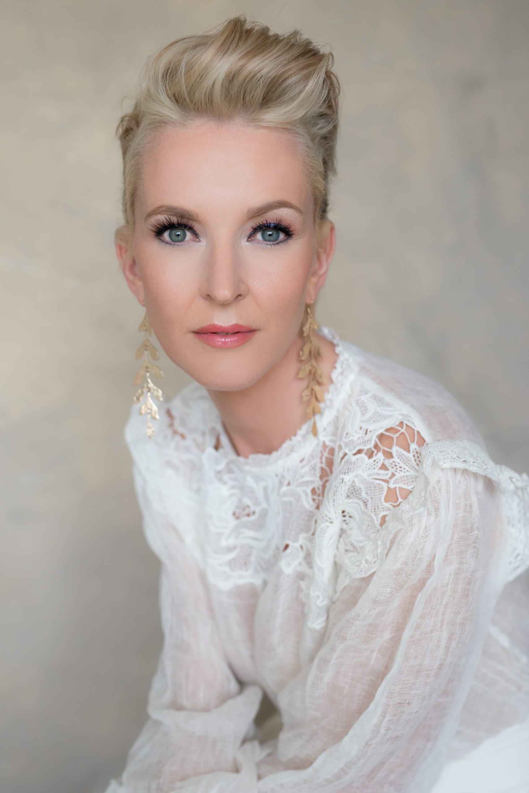 Portrait of a woman with fair skin and blue eyes wearing a sheer white lace blouse and long gold leaf-shaped earrings, with her blonde hair styled back.