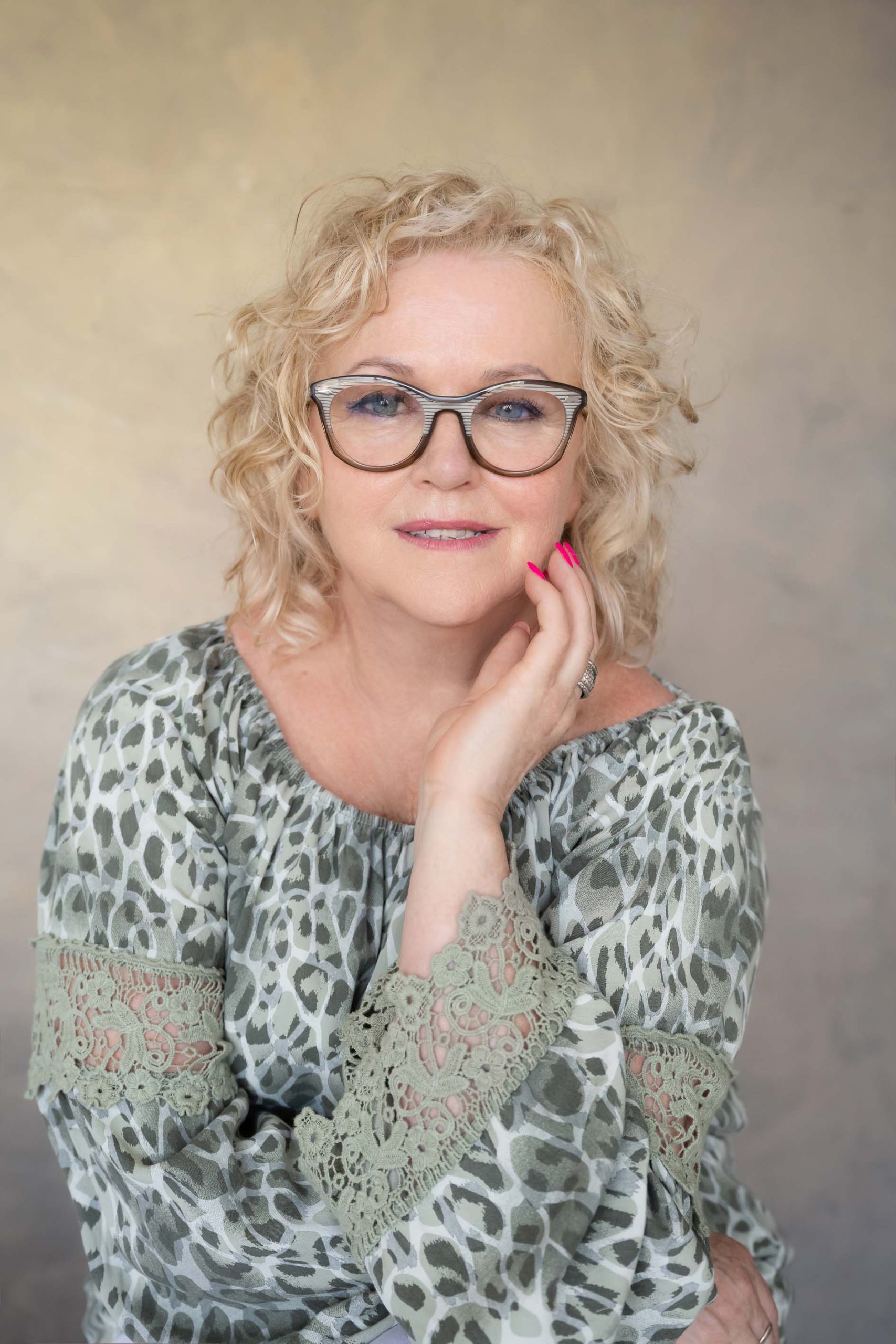 Mature woman with curly blonde hair and glasses wearing a patterned blouse with lace cuffs resting her chin on her hand, smiling gently against a neutral background.