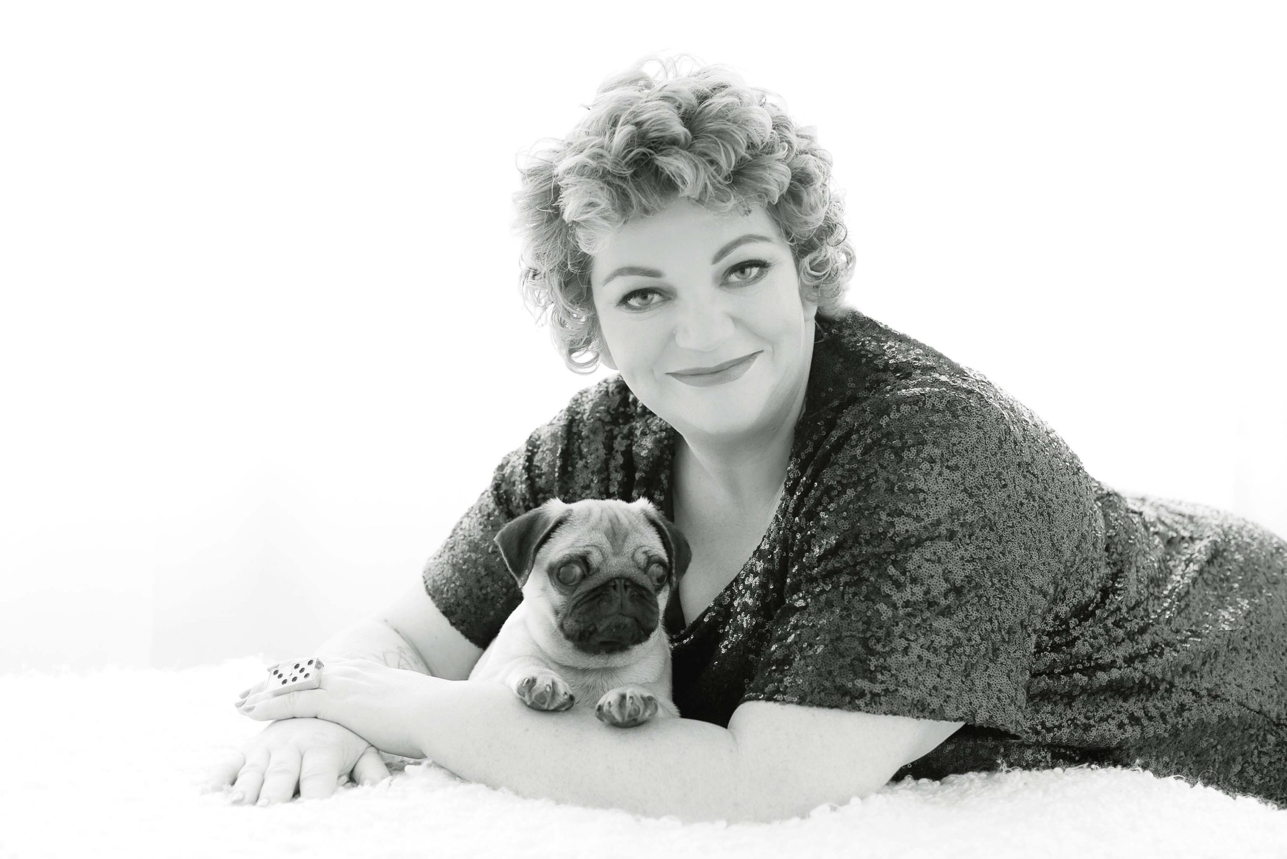 Black and white portrait of a smiling woman with short curly hair lying on a soft surface, wearing a sequined top, holding a small pug dog resting on her arm.