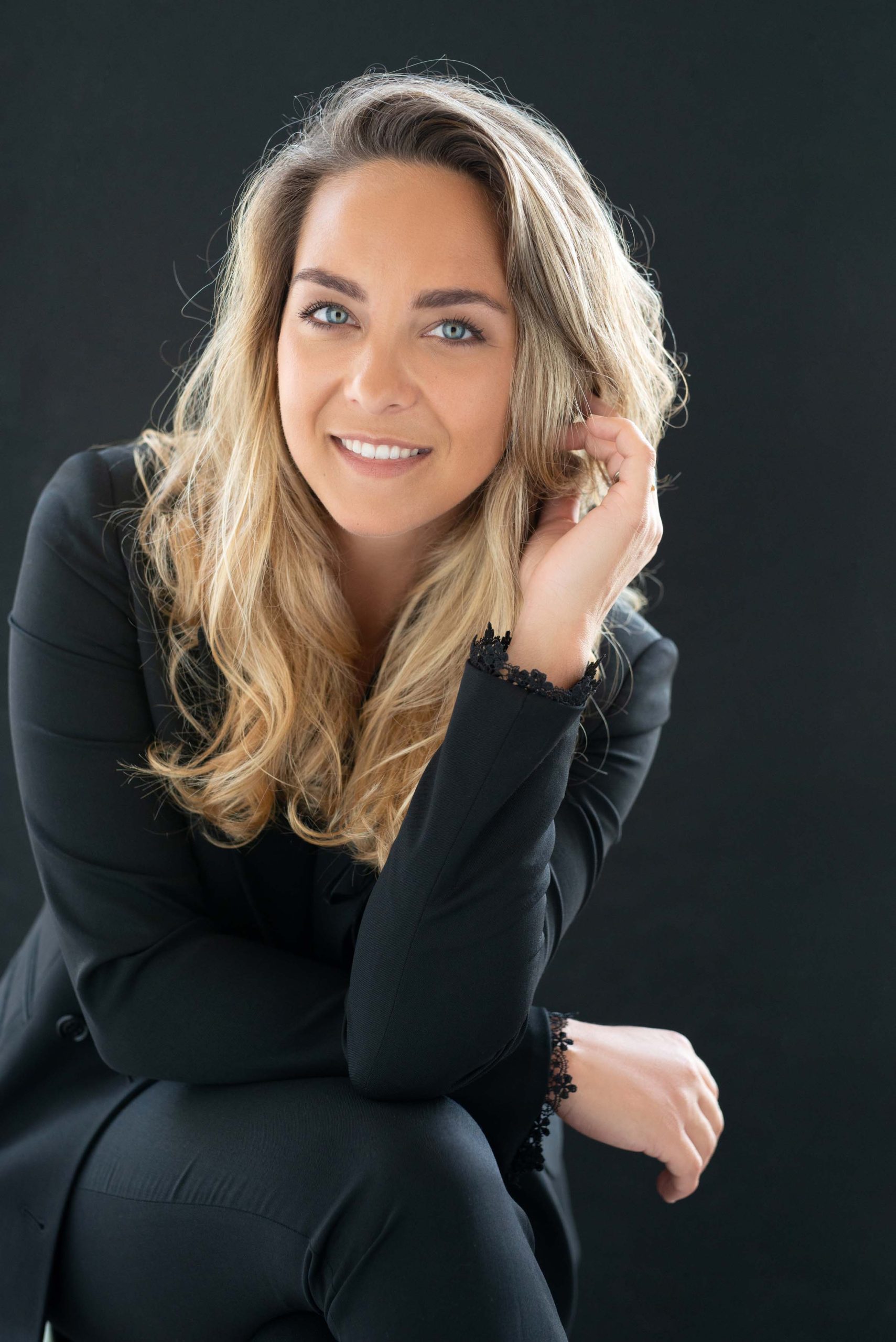 Smiling woman with long blonde hair wearing a black suit jacket sitting against a dark background, resting her chin on her hand.