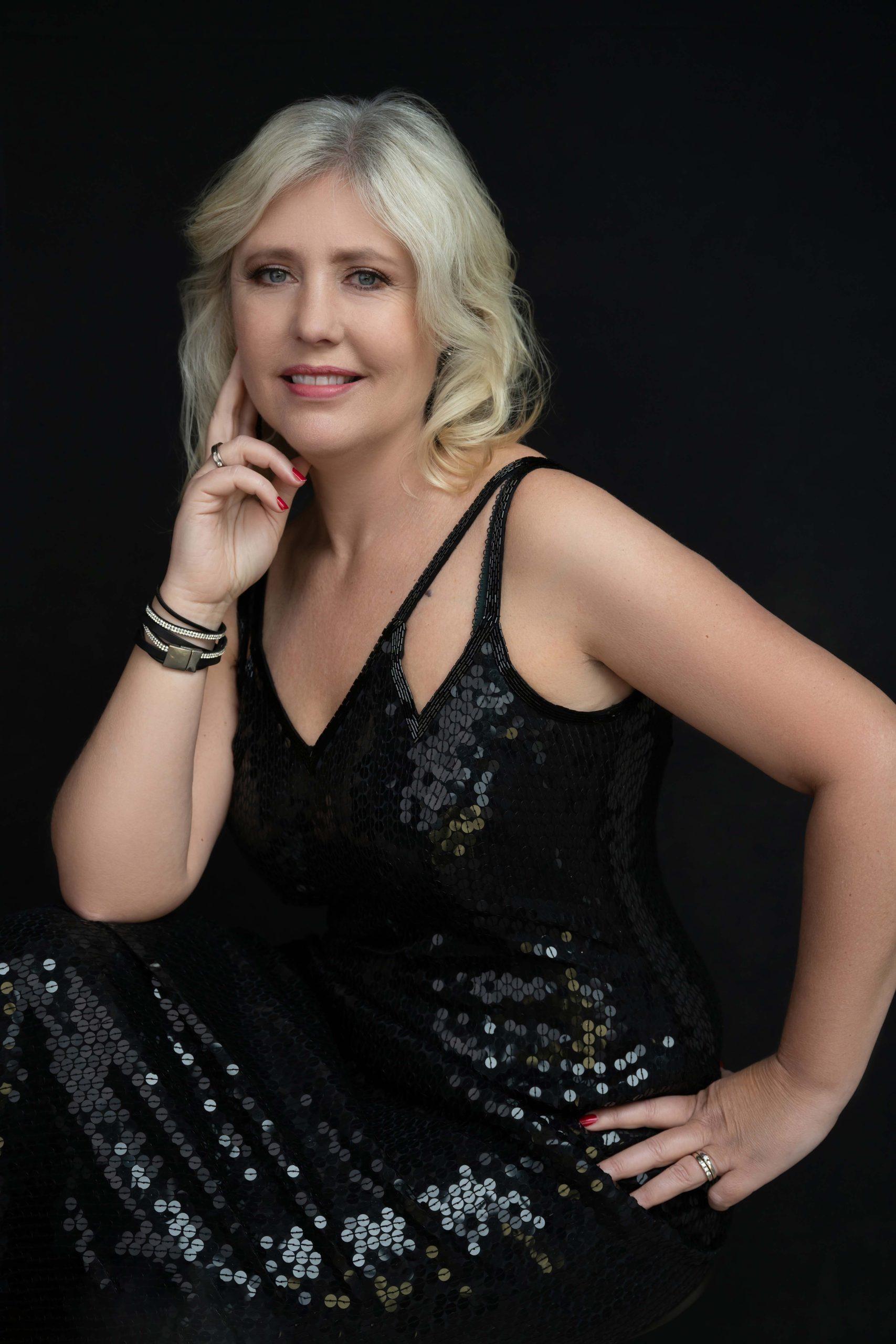 Smiling mature woman with blonde hair wearing a black sequined sleeveless dress, posing with one hand touching her face and the other resting on her hip against a black background.
