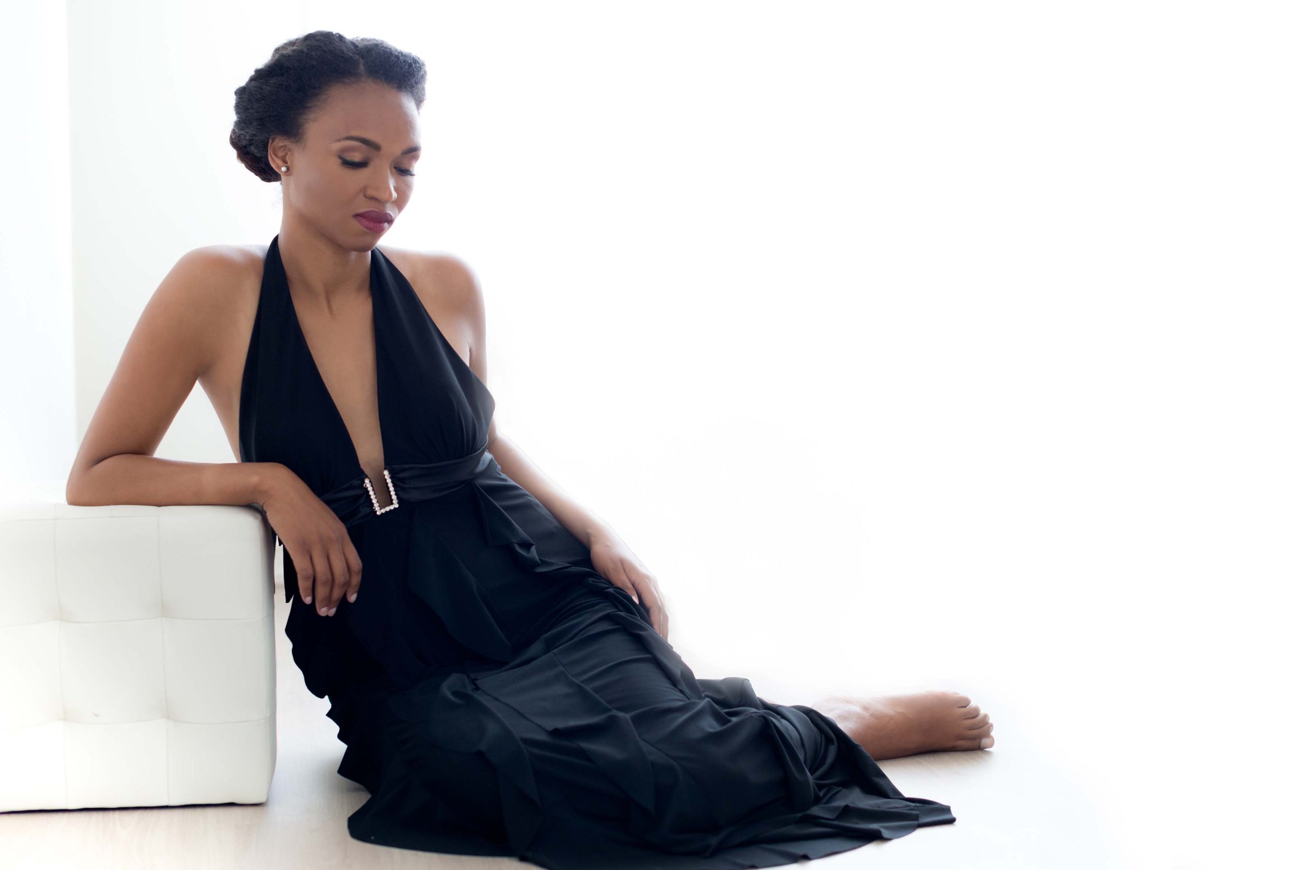 Woman with a stylish updo hairstyle wearing a black halter-neck gown with a jewel belt sitting on the floor and leaning against a white cushioned ottoman, eyes closed.