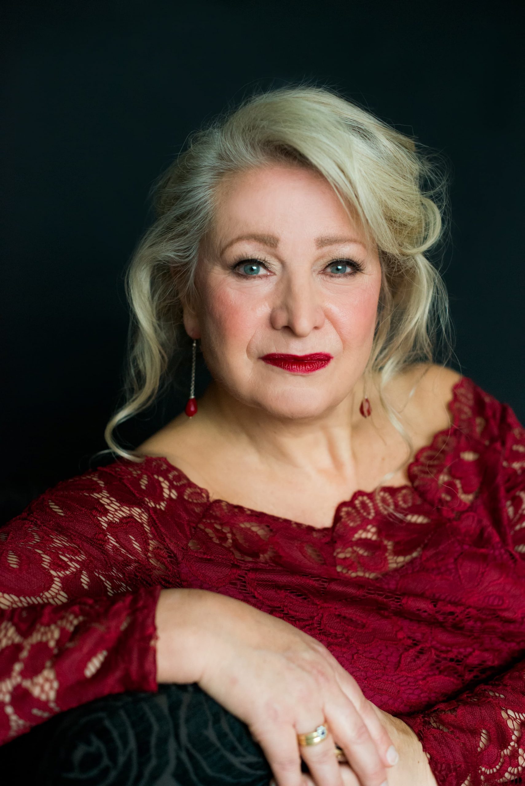 Portrait of an older woman with blonde hair styled in loose waves, wearing a burgundy lace off-shoulder top and red lipstick, posing against a dark background with her hand resting on her knee, showing a gold ring.