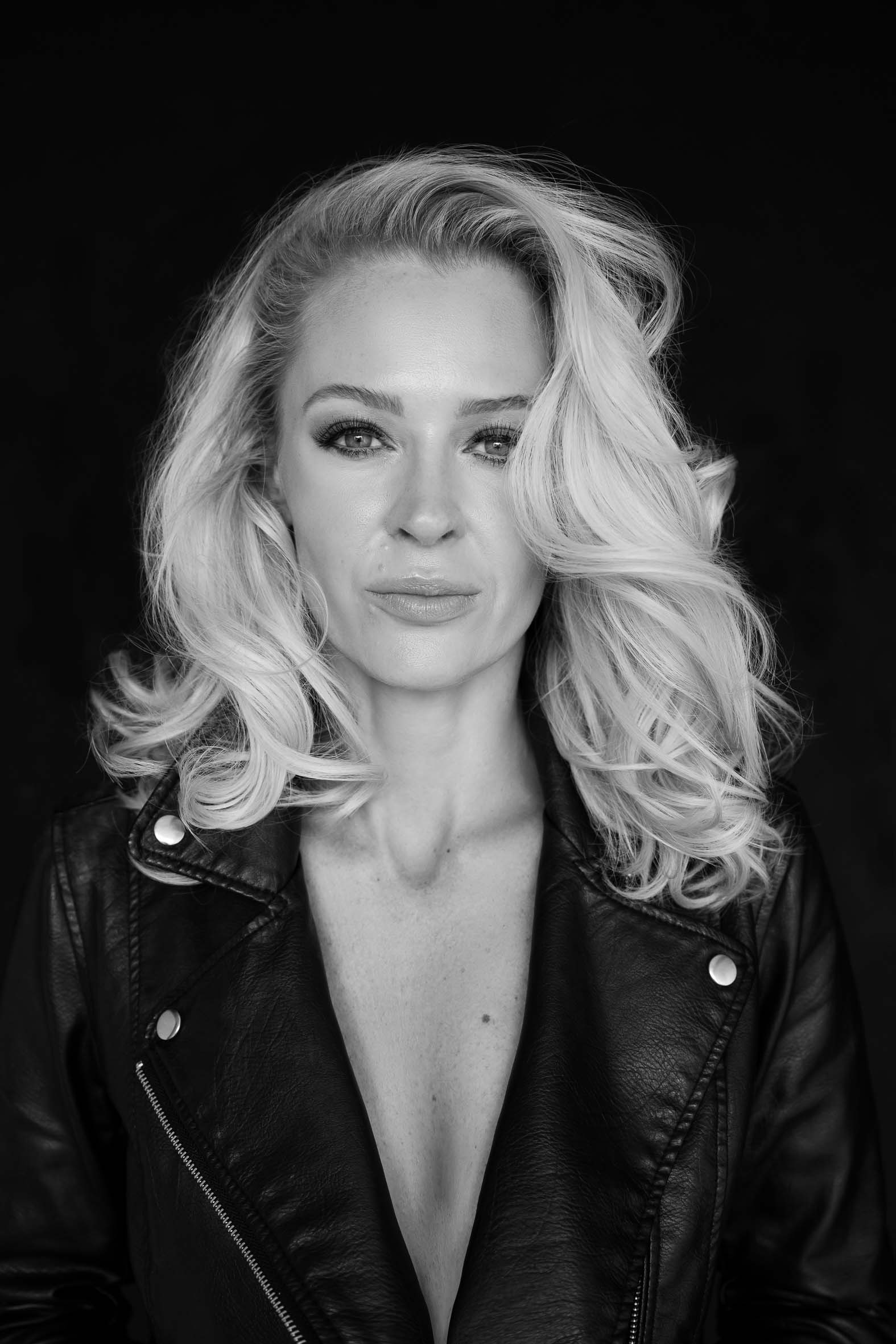 Black and white portrait of a woman with wavy shoulder-length blonde hair wearing a black leather jacket, looking confidently at the camera against a dark background.