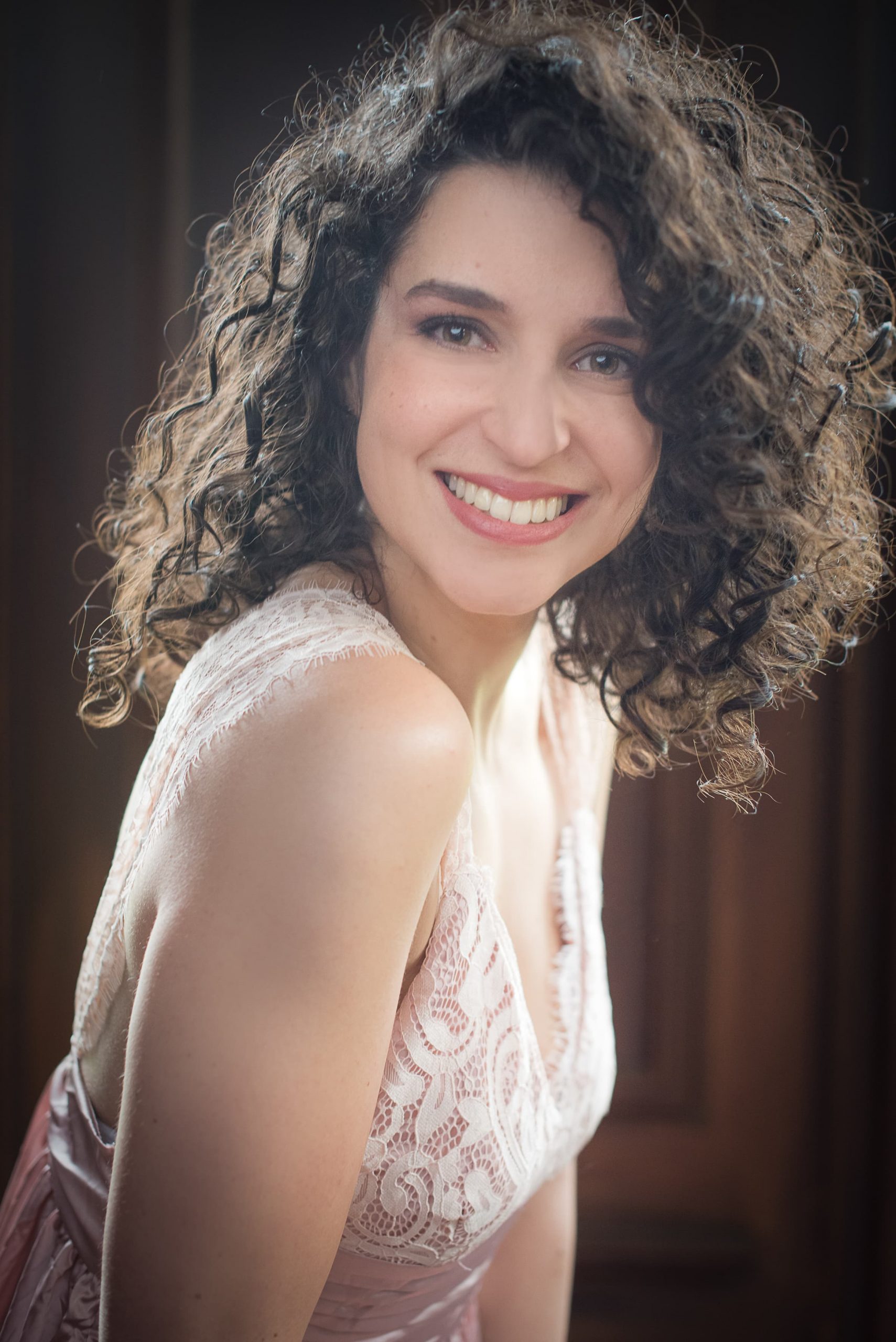 Smiling woman with curly dark hair wearing a pale pink lace dress, posing against a soft-focus brown background.