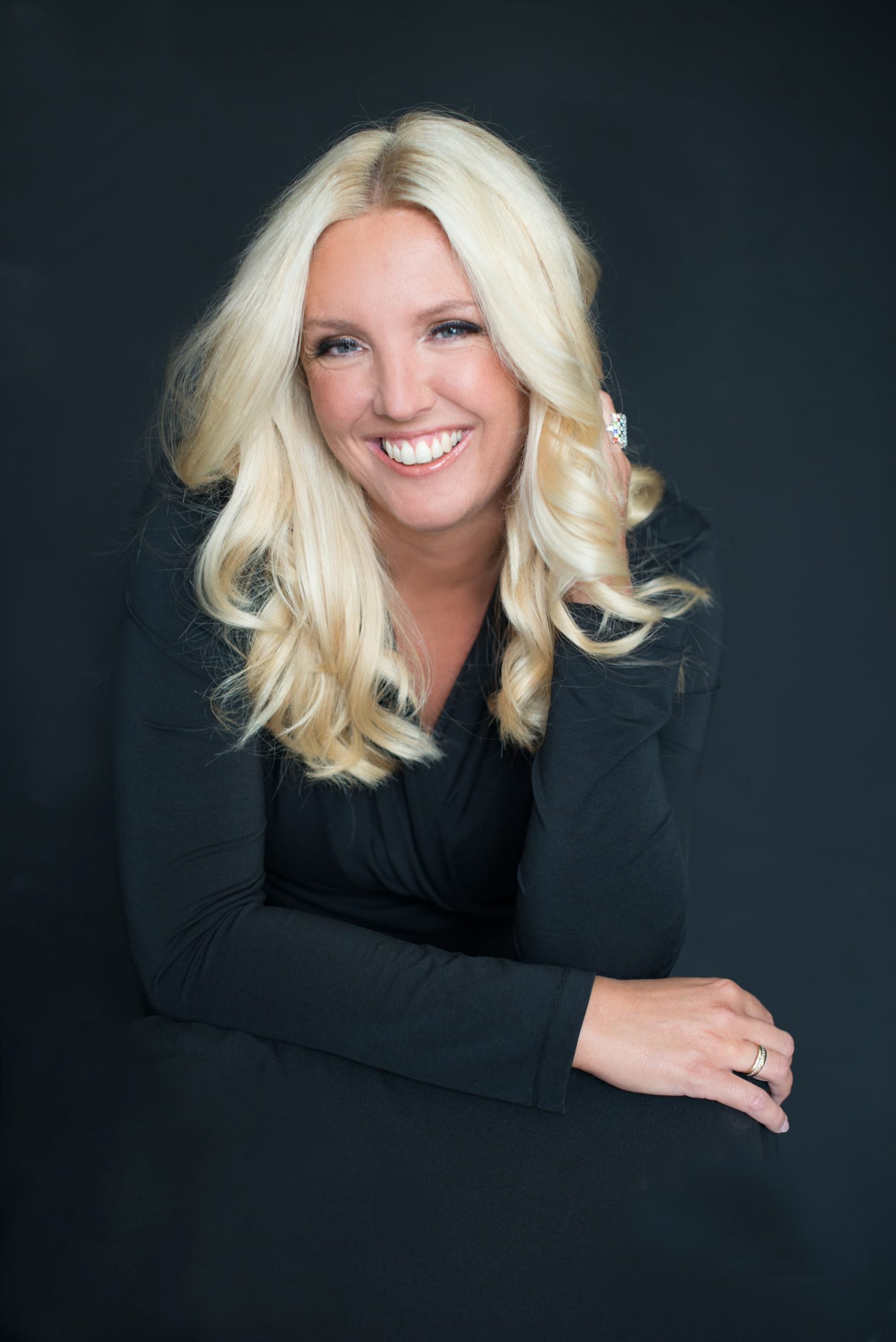 Smiling woman with long curly blonde hair wearing a black long-sleeve top, resting her arms and one hand touching her hair against dark background.