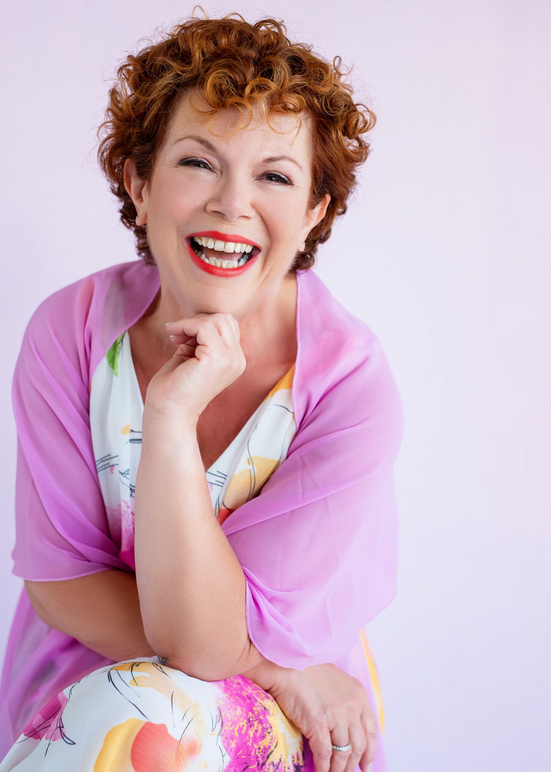Smiling middle-aged woman with curly red hair wearing a colorful dress and a sheer pink shawl, resting her chin on her hand against a light purple background.