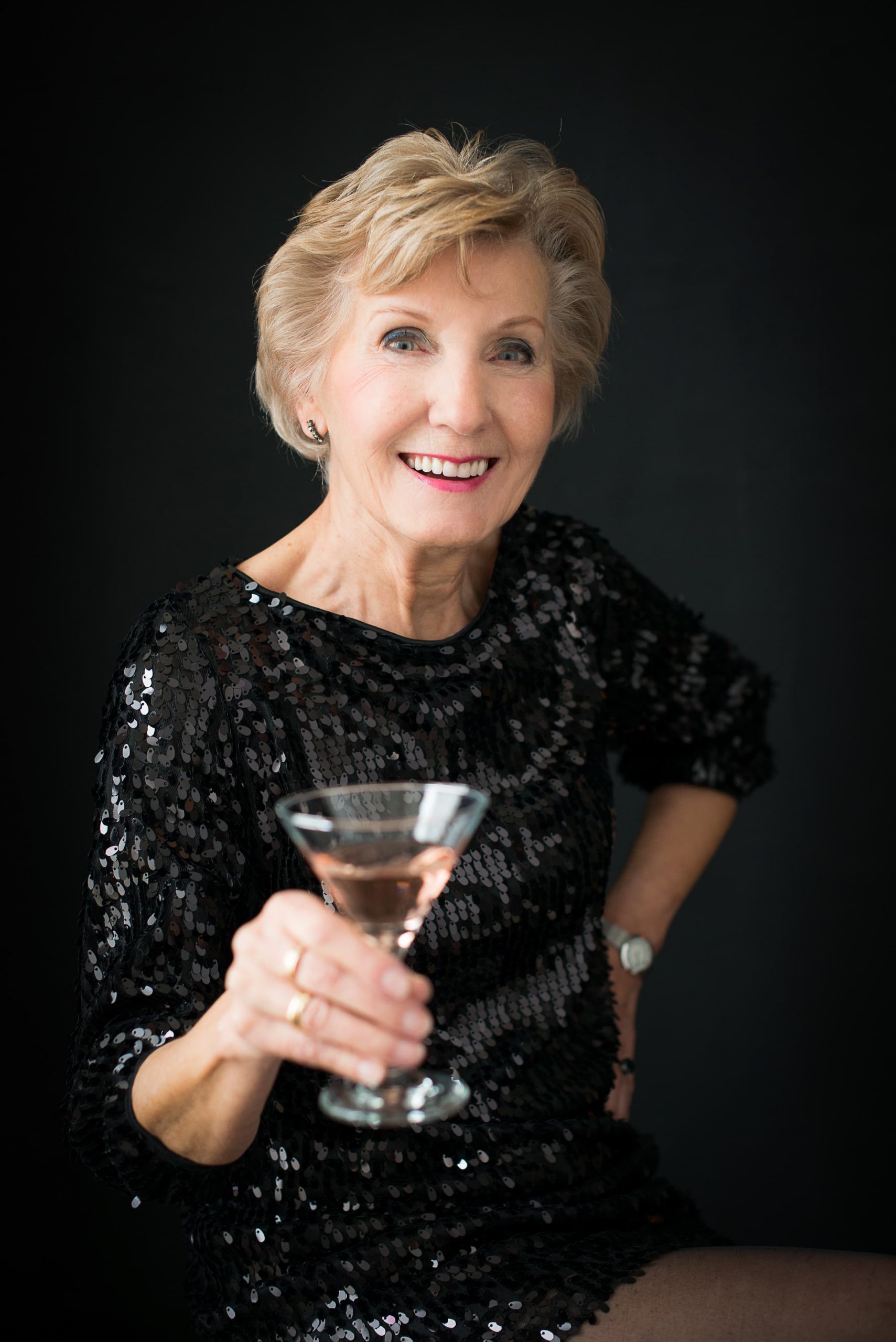 Smiling older woman with short blonde hair wearing a black sequined dress, holding a martini glass with a light pink drink, posed against a black background.