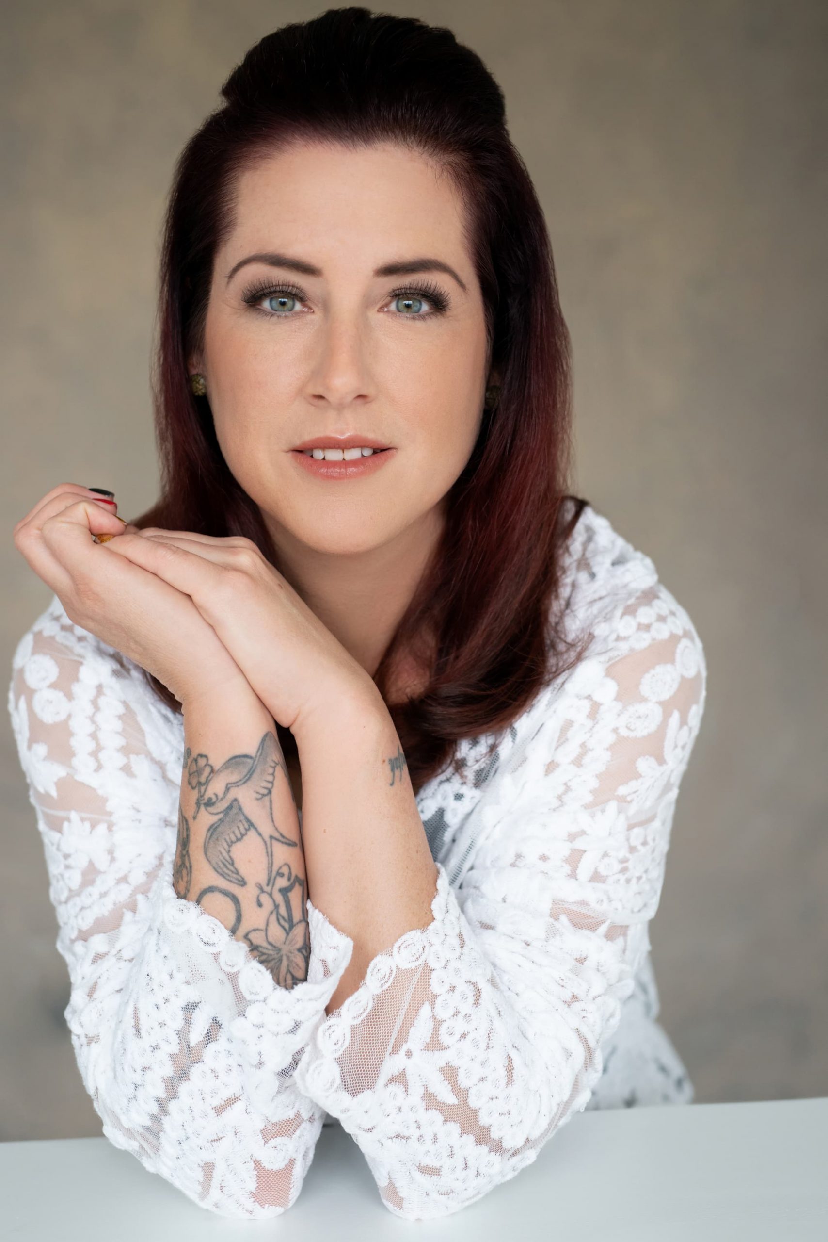 Portrait of a woman with reddish-brown hair and green eyes, wearing a white lace top, resting her crossed tattooed arms on a white surface, looking directly at the camera.
