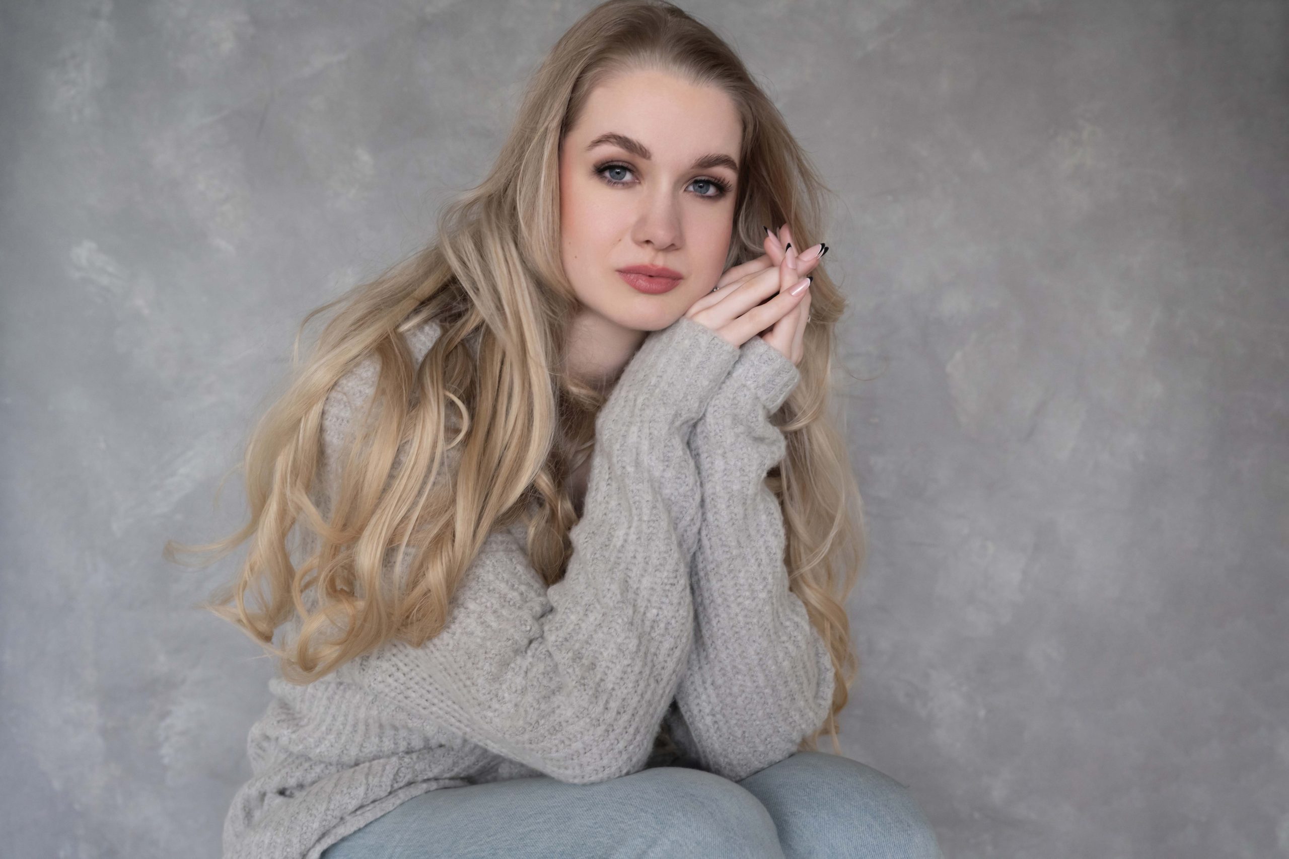 Young woman with long blonde hair wearing a gray knitted sweater, sitting with hands clasped near her face against a textured gray background.