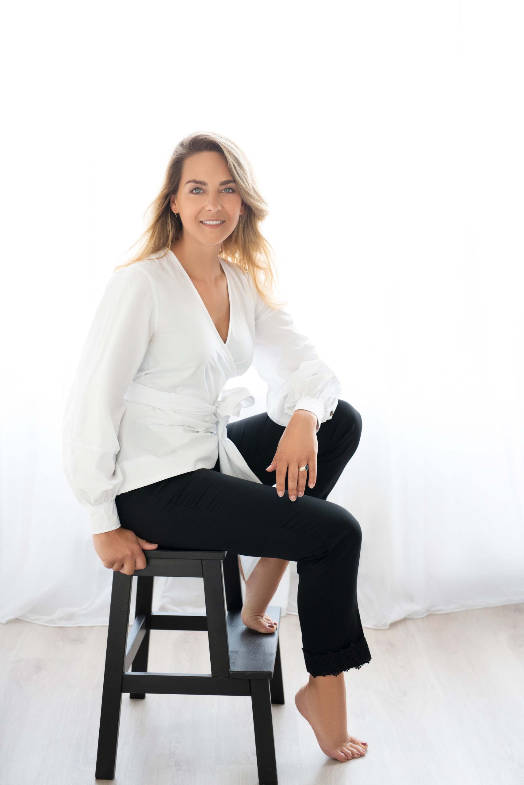 A woman with blonde hair wearing a white wrap blouse and black pants sits barefoot on a black wooden stool against a bright white background, smiling gently.