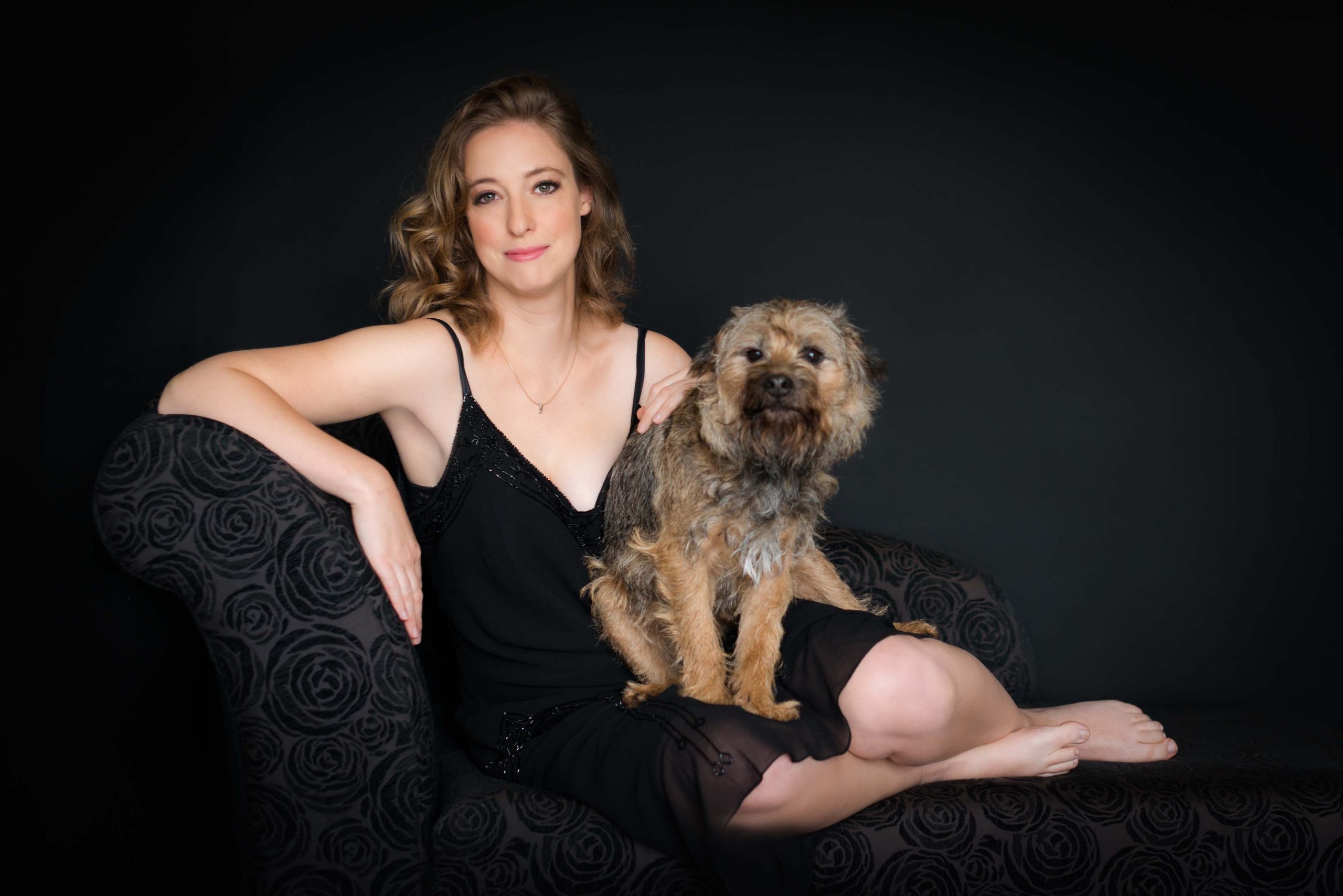 Woman in a black dress sitting barefoot on a black floral patterned sofa with a small brown dog on her lap against a dark background.
