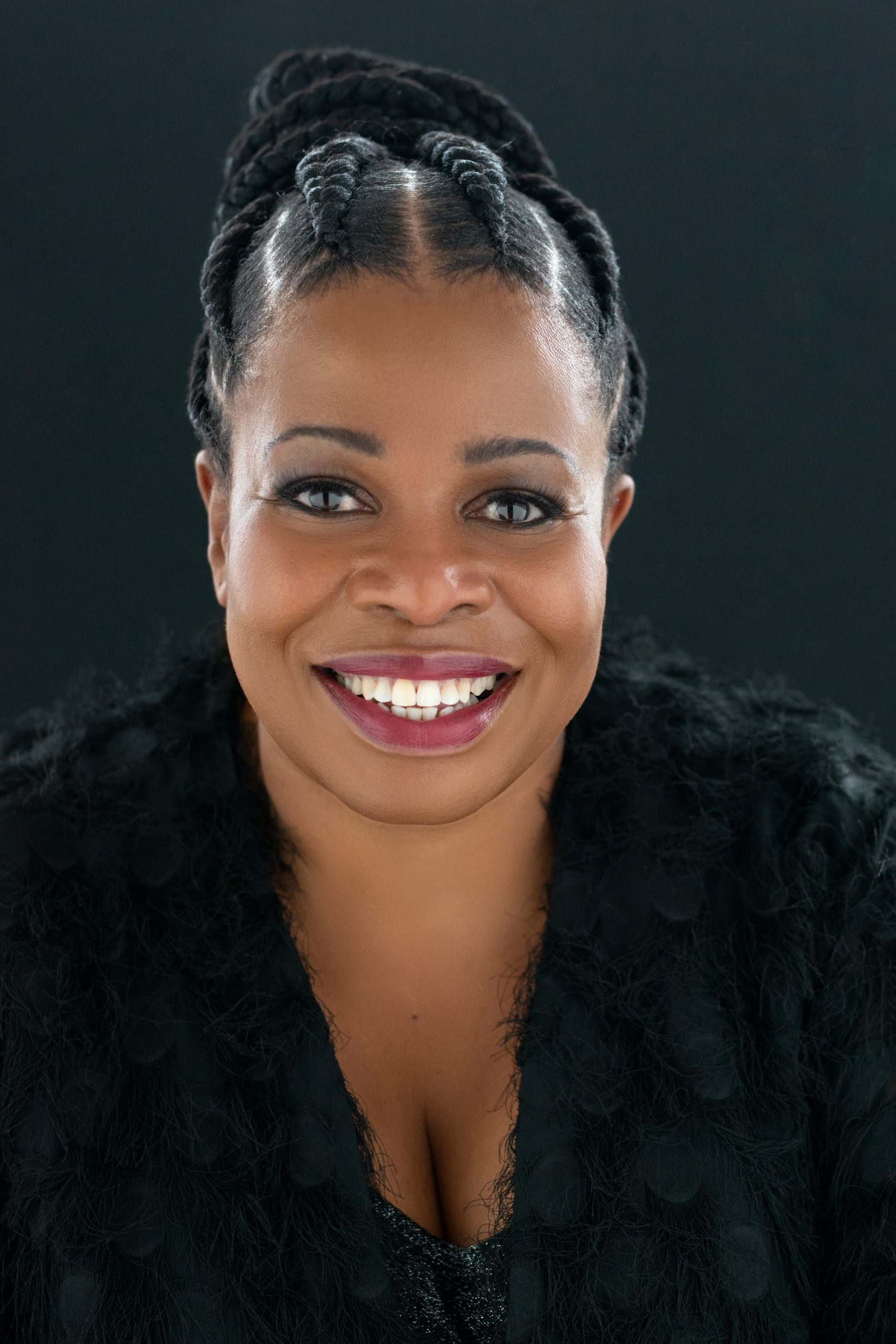 Portrait of a smiling Black woman with braided hair styled upwards, wearing dark eye makeup and purple lipstick, dressed in a textured black garment, against a dark background.
