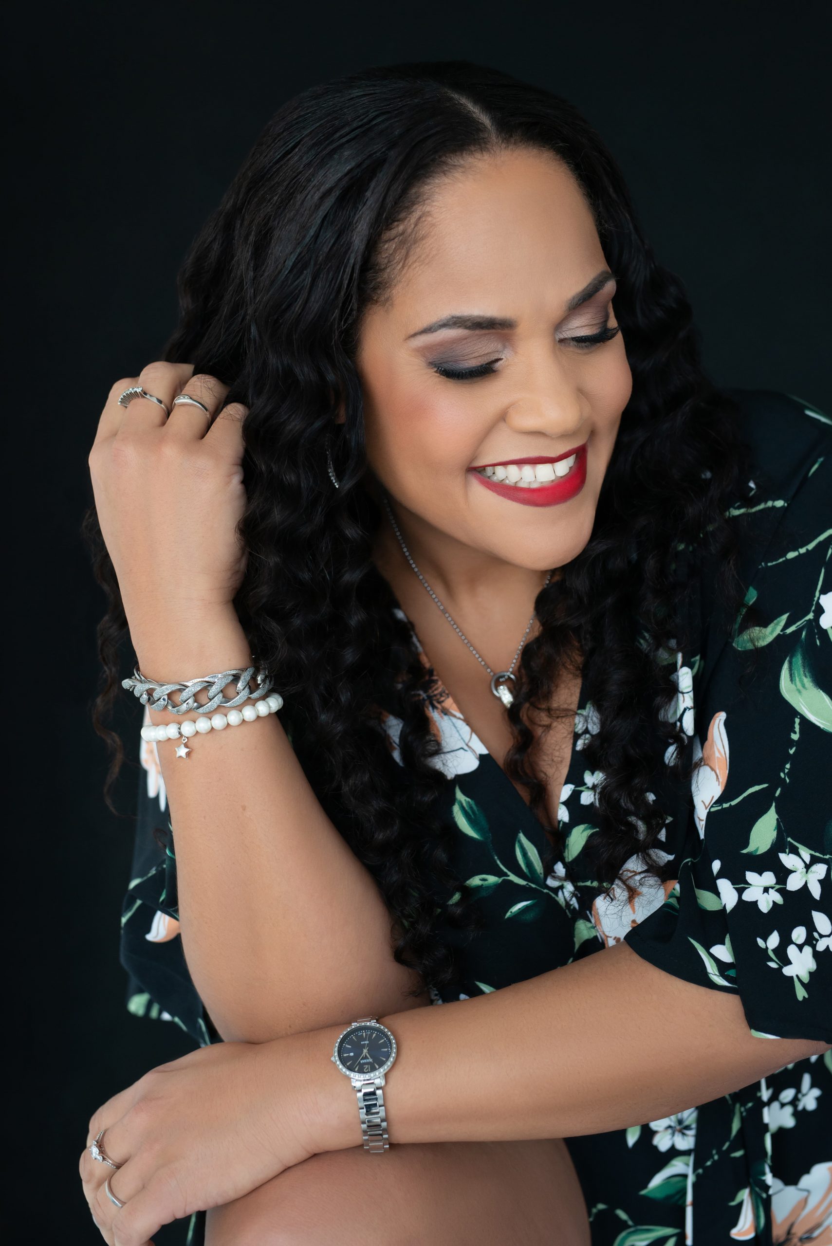 Smiling woman with long curly black hair wearing a black floral dress, silver watch, and bracelets, sitting against a dark background with eyes closed and a joyful expression.