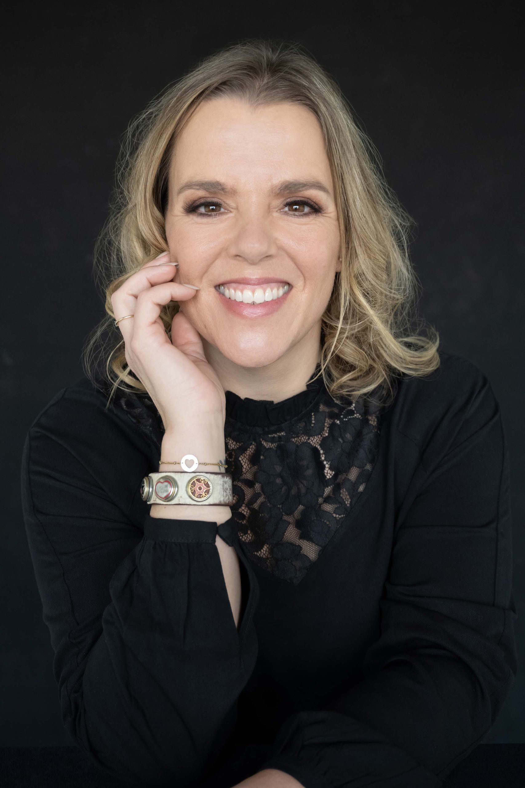 Smiling woman with shoulder-length blonde hair wearing a black lace top and bracelets, posing against a black background with her hand touching her face.