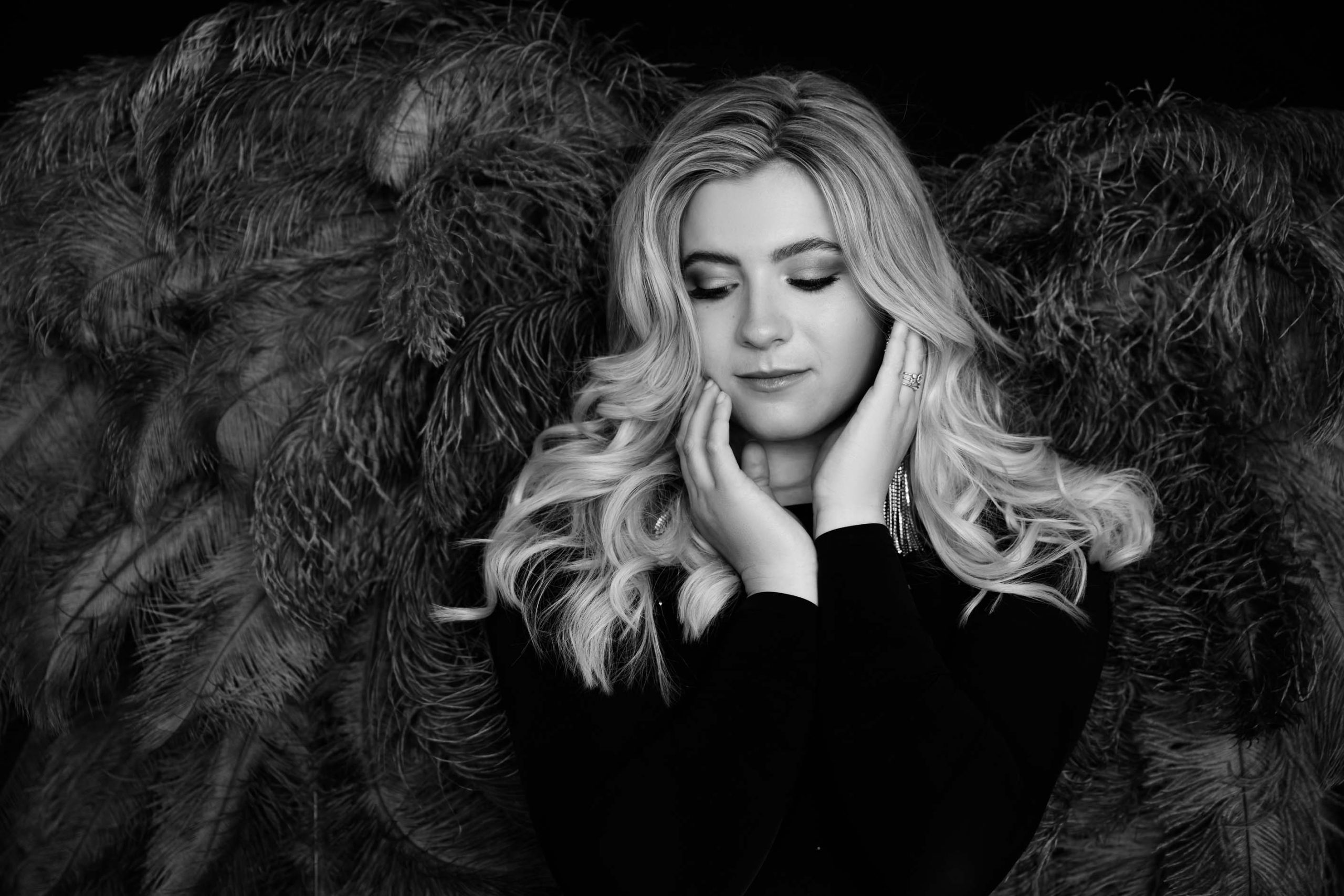 Black and white portrait of a woman with long wavy blonde hair, wearing a dark top with earrings and a ring, gently touching her face with closed eyes and a peaceful expression, against a backdrop of textured feathers.