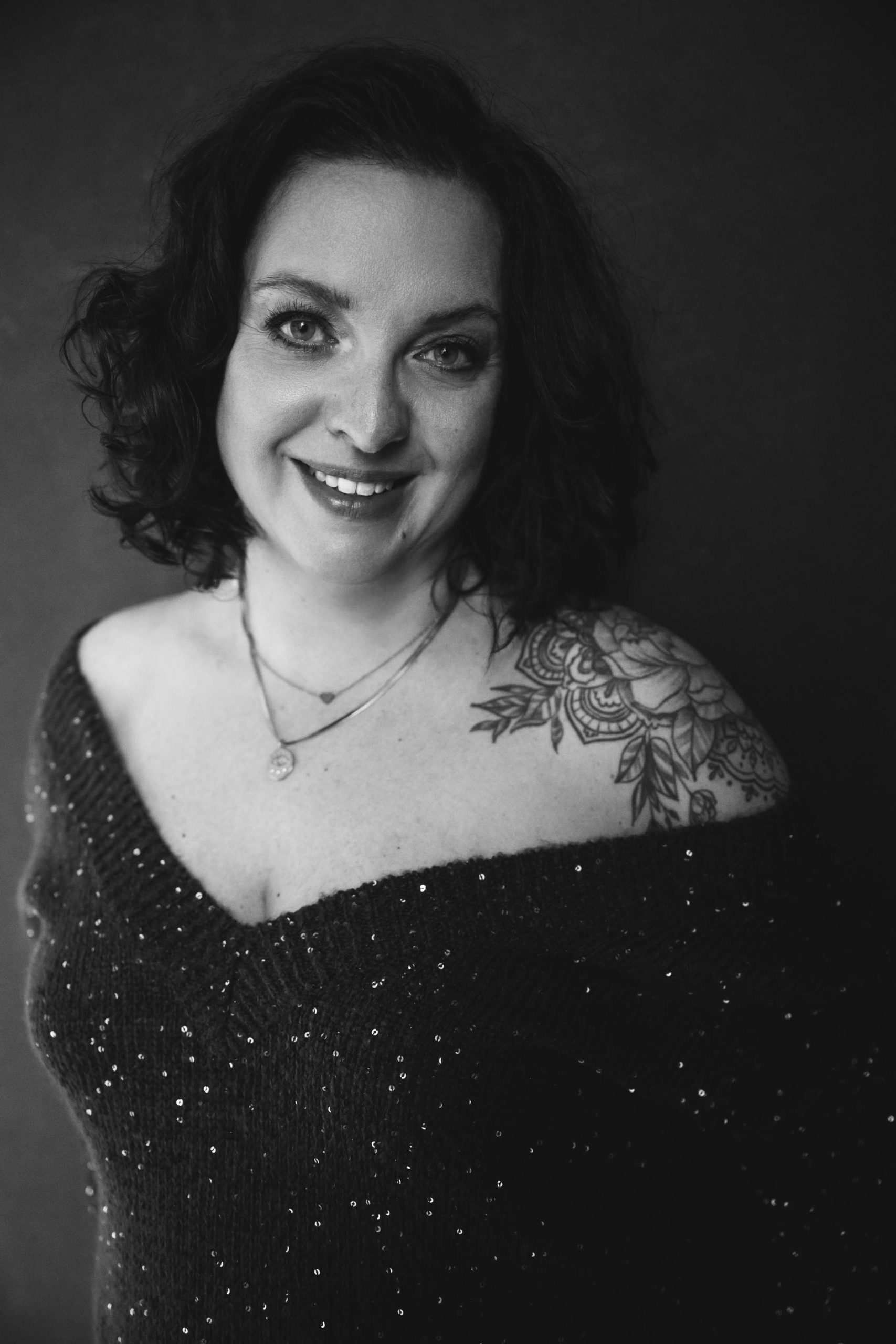 Black and white portrait of a smiling woman with short curly hair wearing a dark off-shoulder sweater adorned with small sequins, showing a floral tattoo on her left shoulder and two delicate necklaces.